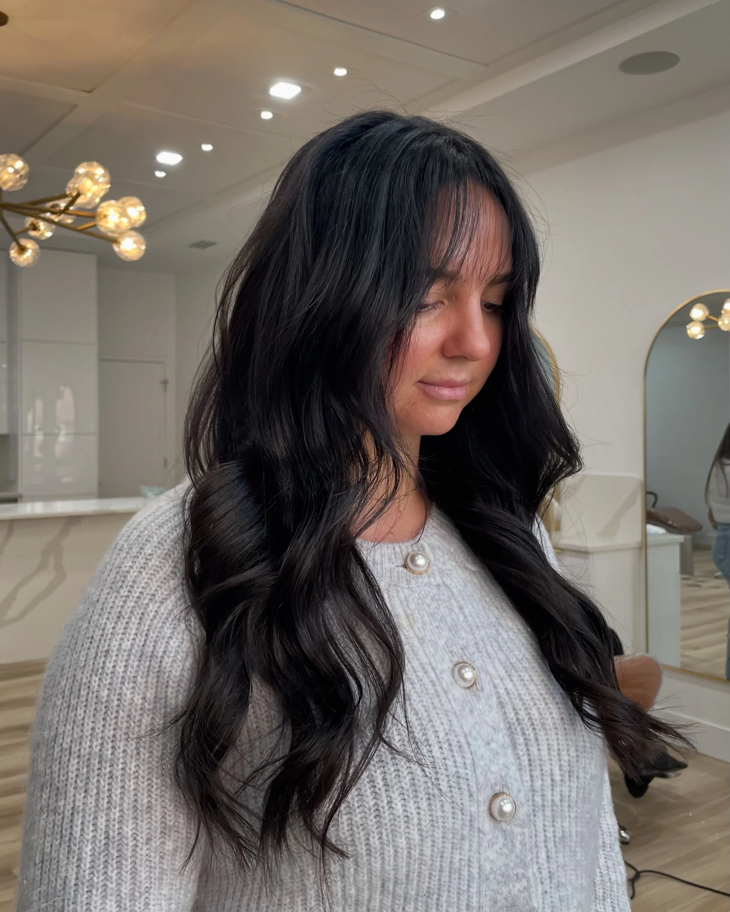 Woman with long black hair in loose curls, wearing a light-colored sweater with pearl buttons, standing in a modern, well-lit space with decorative wall mirror and contemporary light fixtures.