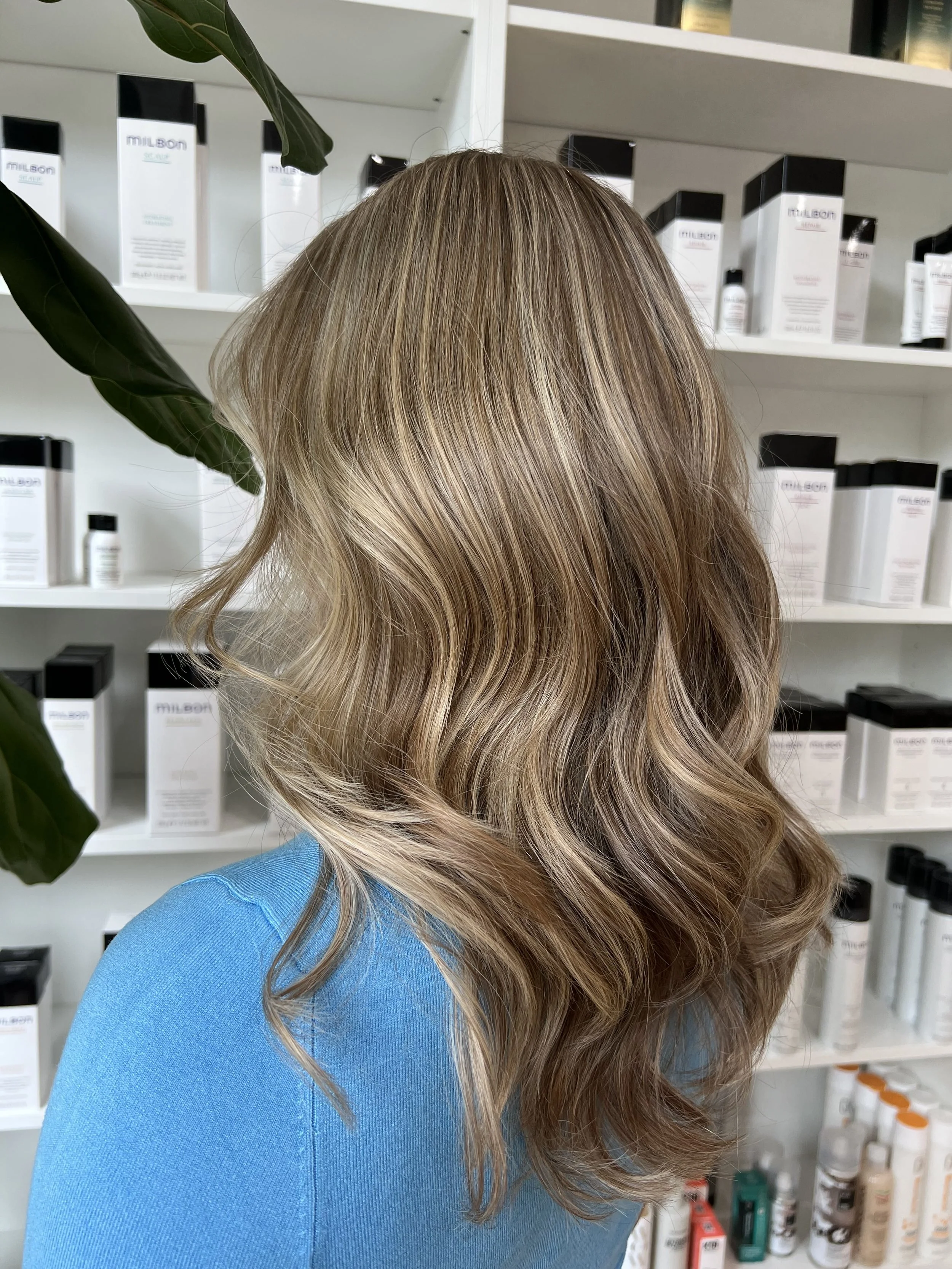 A woman with wavy blonde hair wearing a blue top standing in front of shelves filled with skincare products.