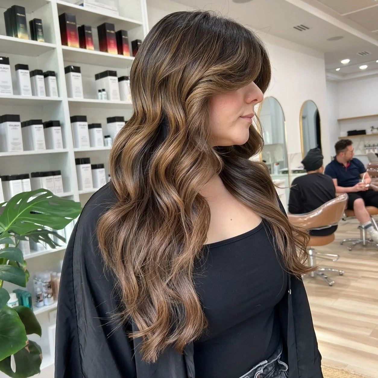 Woman with long, wavy brown hair in a salon, wearing a black top, with hair products in the background.