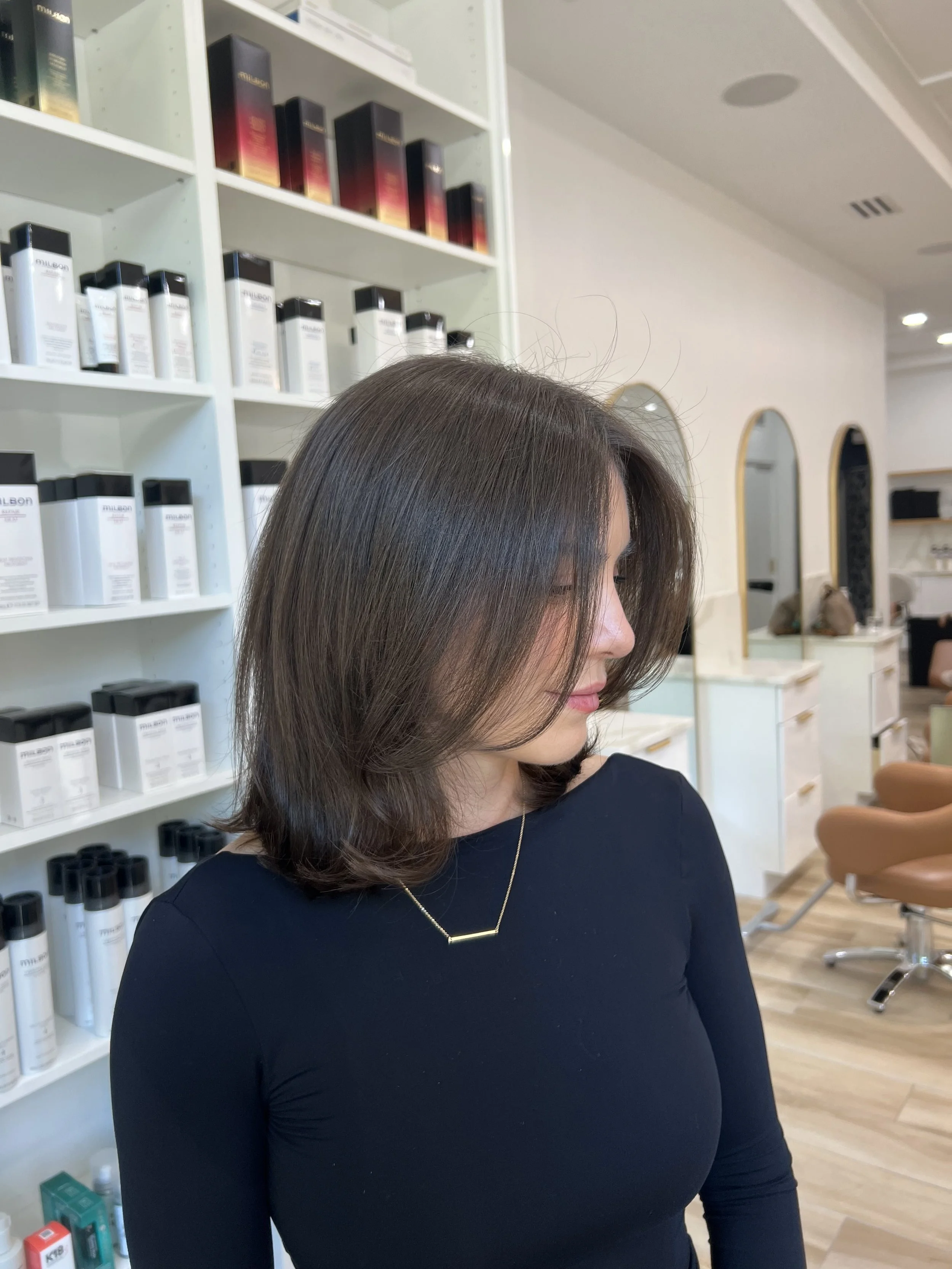 A woman with shoulder-length brown hair wearing a black top and a gold necklace with a bar pendant, sitting in a salon or spa setting.
