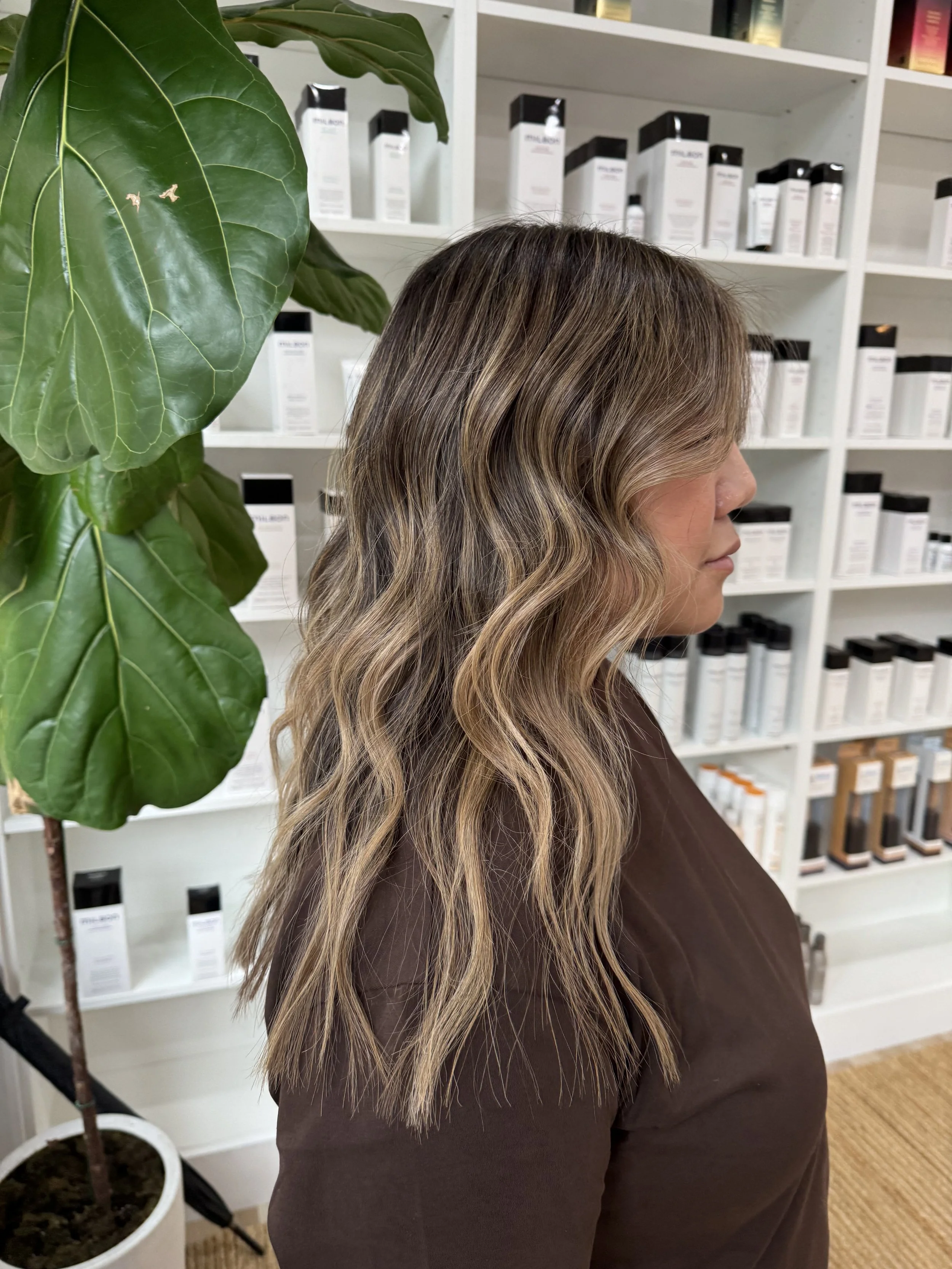 A woman with wavy, blonde-brown hair standing in front of a white bookshelf filled with skincare products, next to a large green plant.