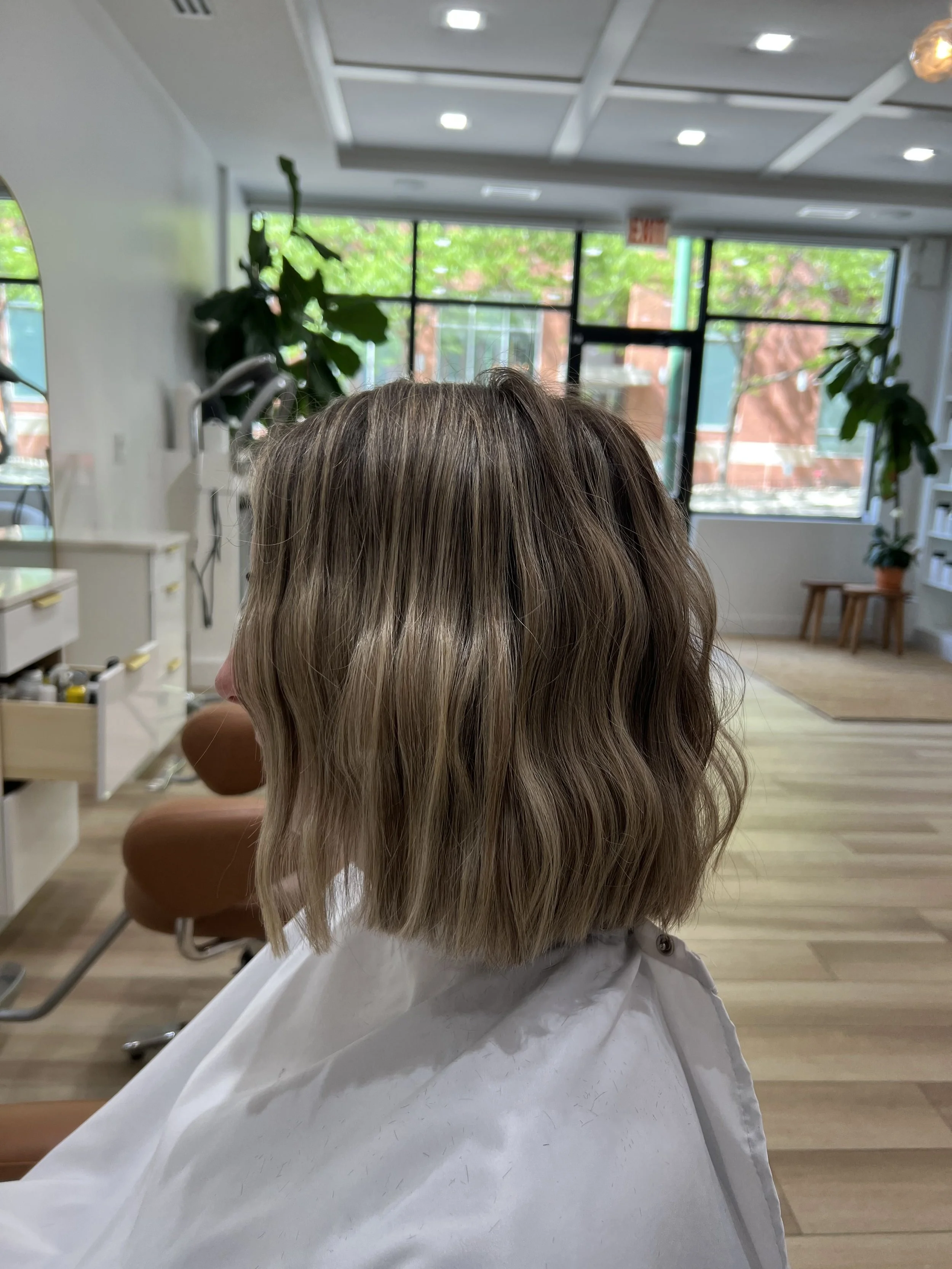 Person with shoulder-length wavy blonde hair sitting in a salon chair with a white cape, inside a modern hair salon with large windows and indoor plants.