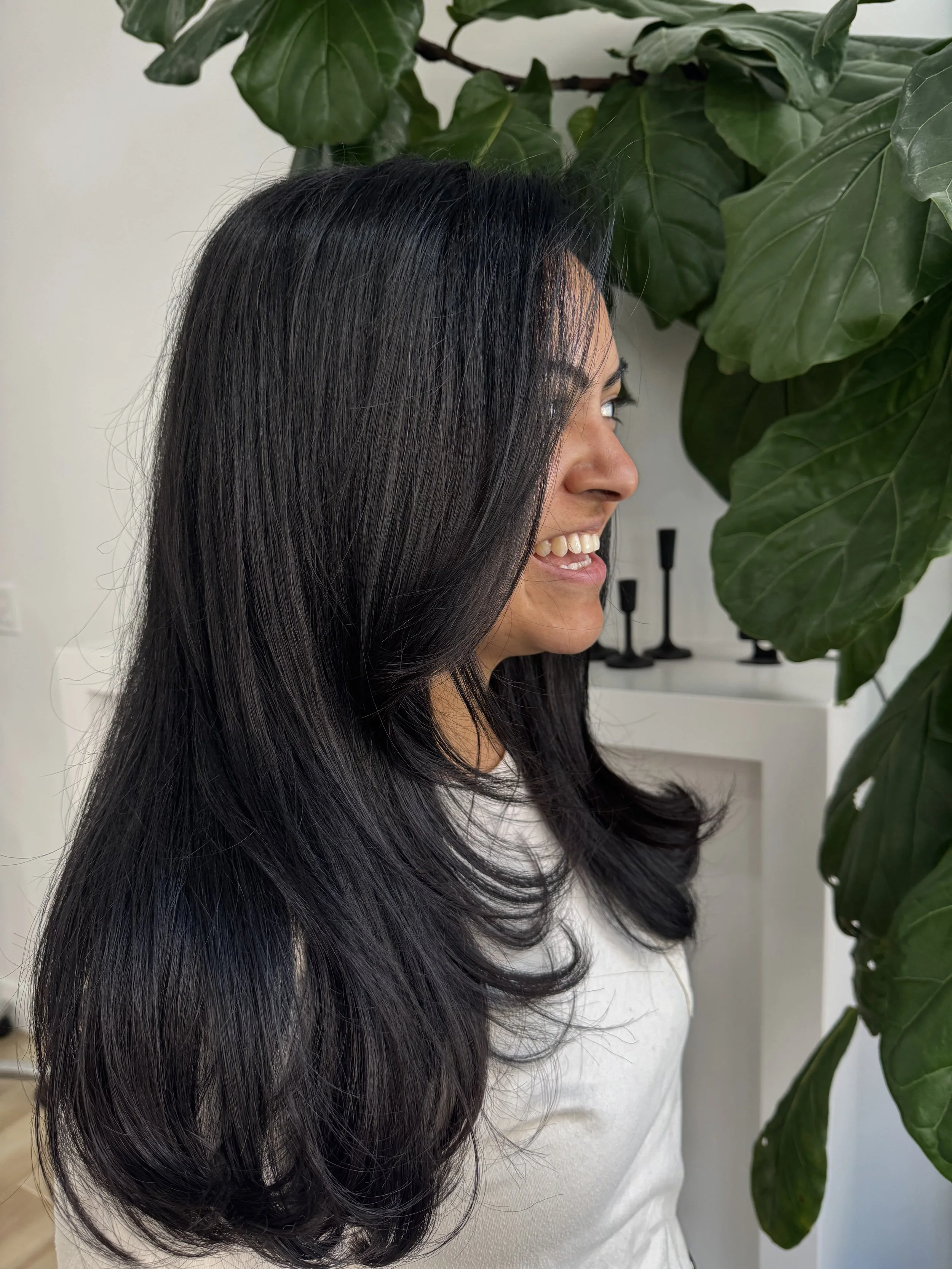 A woman with long black hair smiling and looking to her right, standing next to large green indoor leaves.