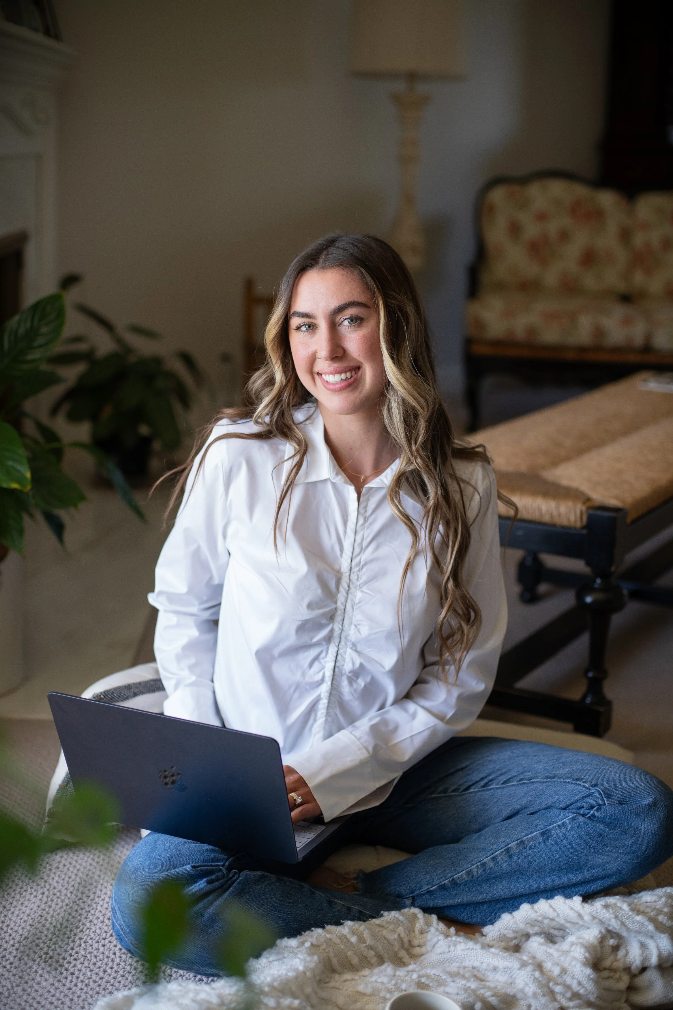 A young woman with long blonde wavy hair sitting cross-legged indoors, holding a laptop on her lap, smiling at the camera.