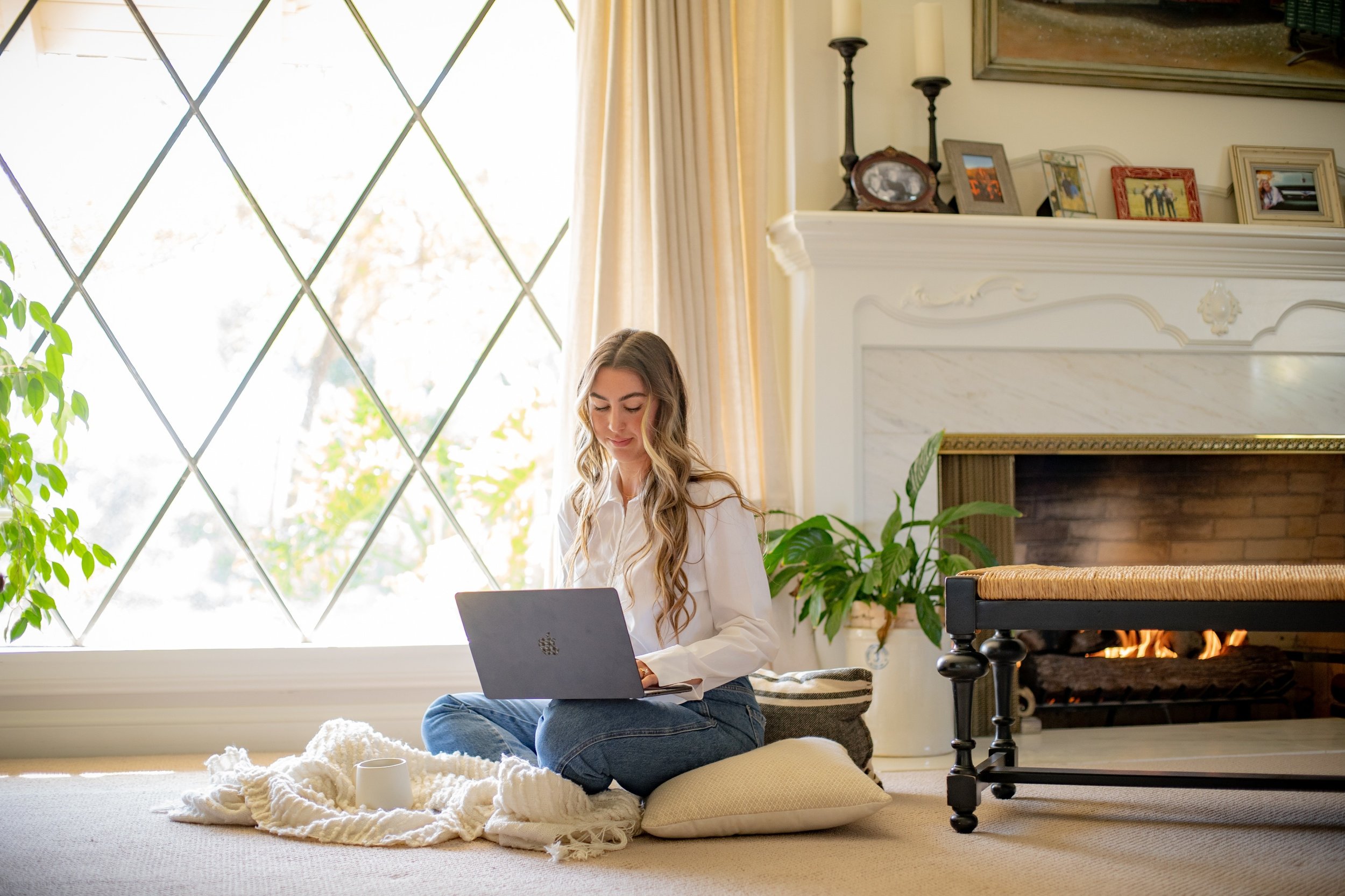 A young woman with long blonde hair, wearing a white shirt and jeans, sitting cross-legged on a cushion on a beige carpet in front of a large window with curtain, working on a laptop. The room has a white fireplace with framed photos and candlesticks on the mantel, a potted plant, and a cozy setting with natural light.