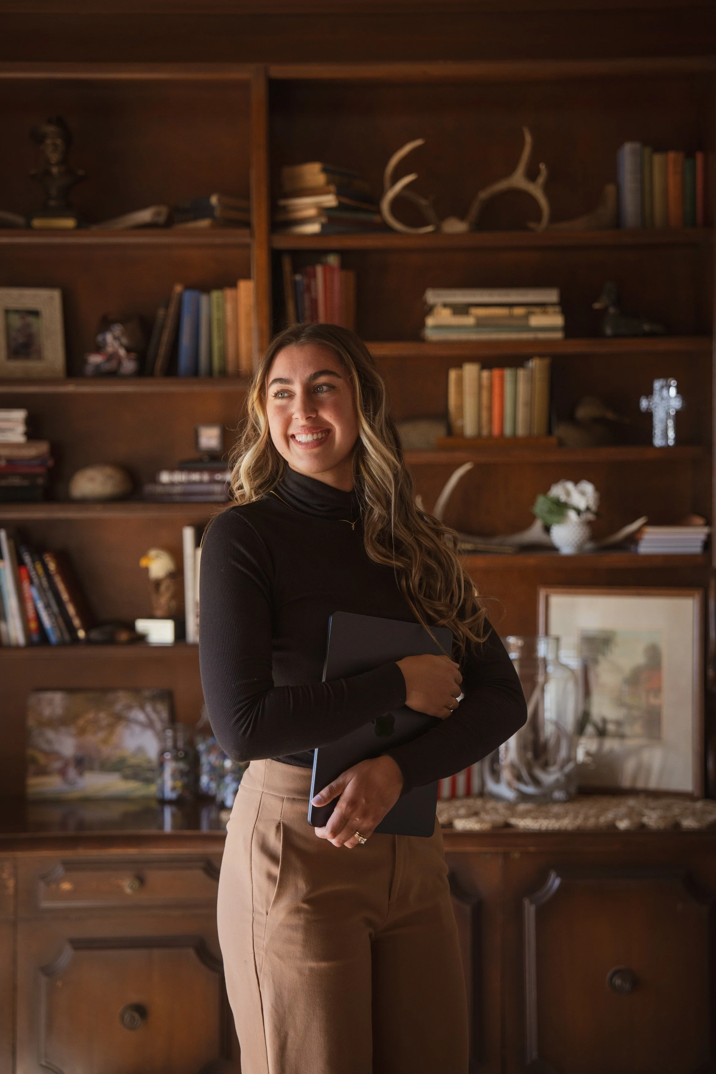 A smiling woman with long wavy hair, wearing a black turtleneck and tan pants, holding a closed laptop in front of a wooden bookshelf filled with books, decorative items, and a cross