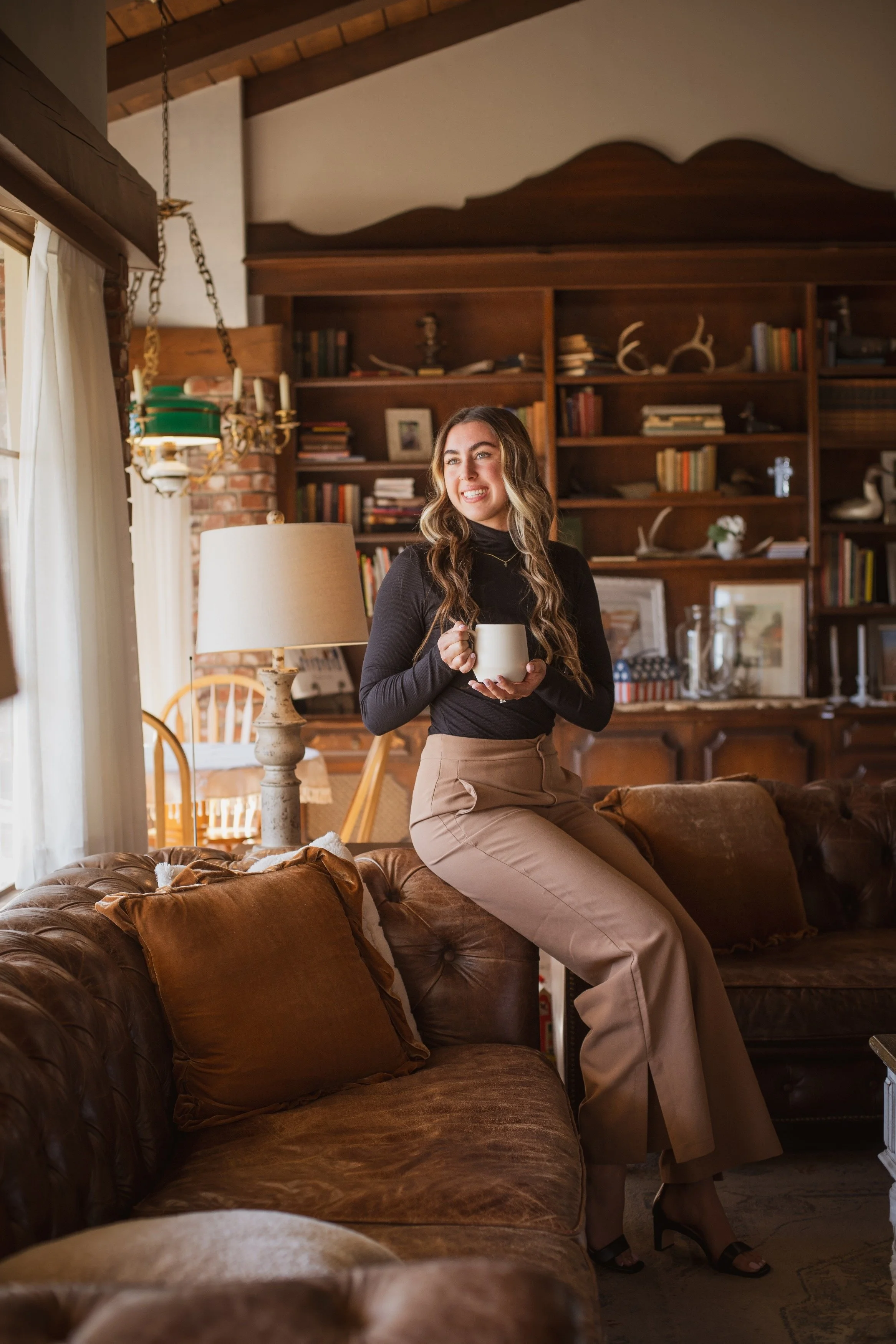 A woman with long, wavy hair wearing a black turtleneck and beige pants, sitting on the arm of a leather sofa, holding a coffee mug, in a cozy, well-decorated living room with bookshelves, a lamp, and natural light.