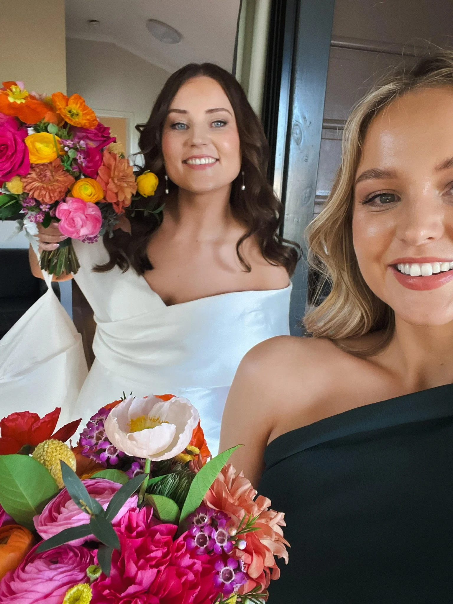 Two women smiling, one holding a bouquet of vibrant pink, yellow, orange, and purple flowers, with one woman wearing a white off-shoulder dress and the other in a black off-shoulder dress, indoors near an open door.