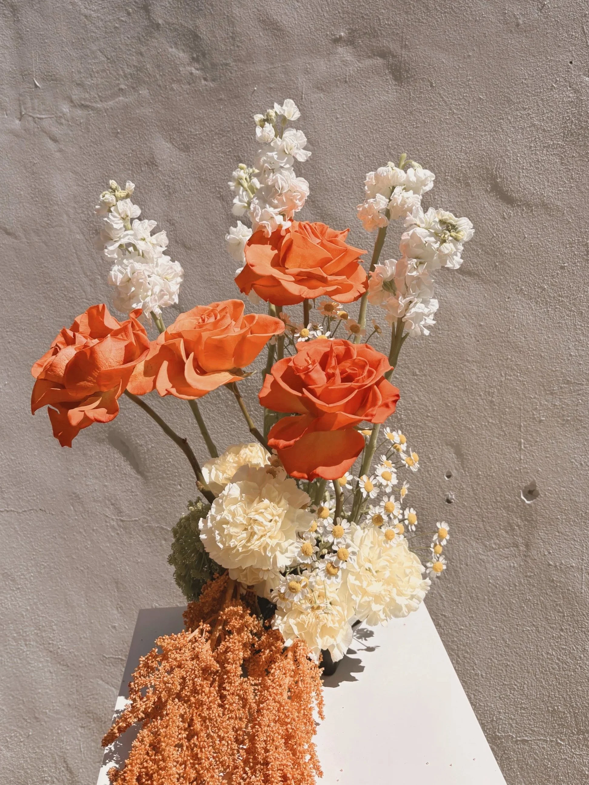 A bouquet of orange roses, white carnations, white daisies, and peach-colored pampas grass arranged on a white surface against a textured beige wall, with a clear blue sky and trees in the background.