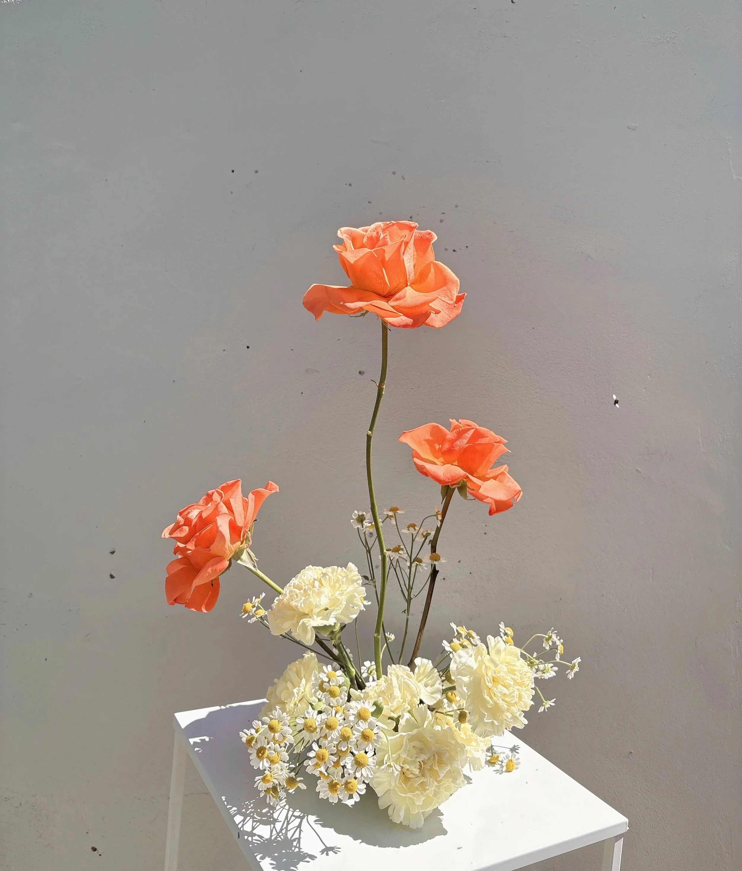 A floral arrangement with peach roses, white carnations, and daisies on a white table against a plain wall with sunlight.