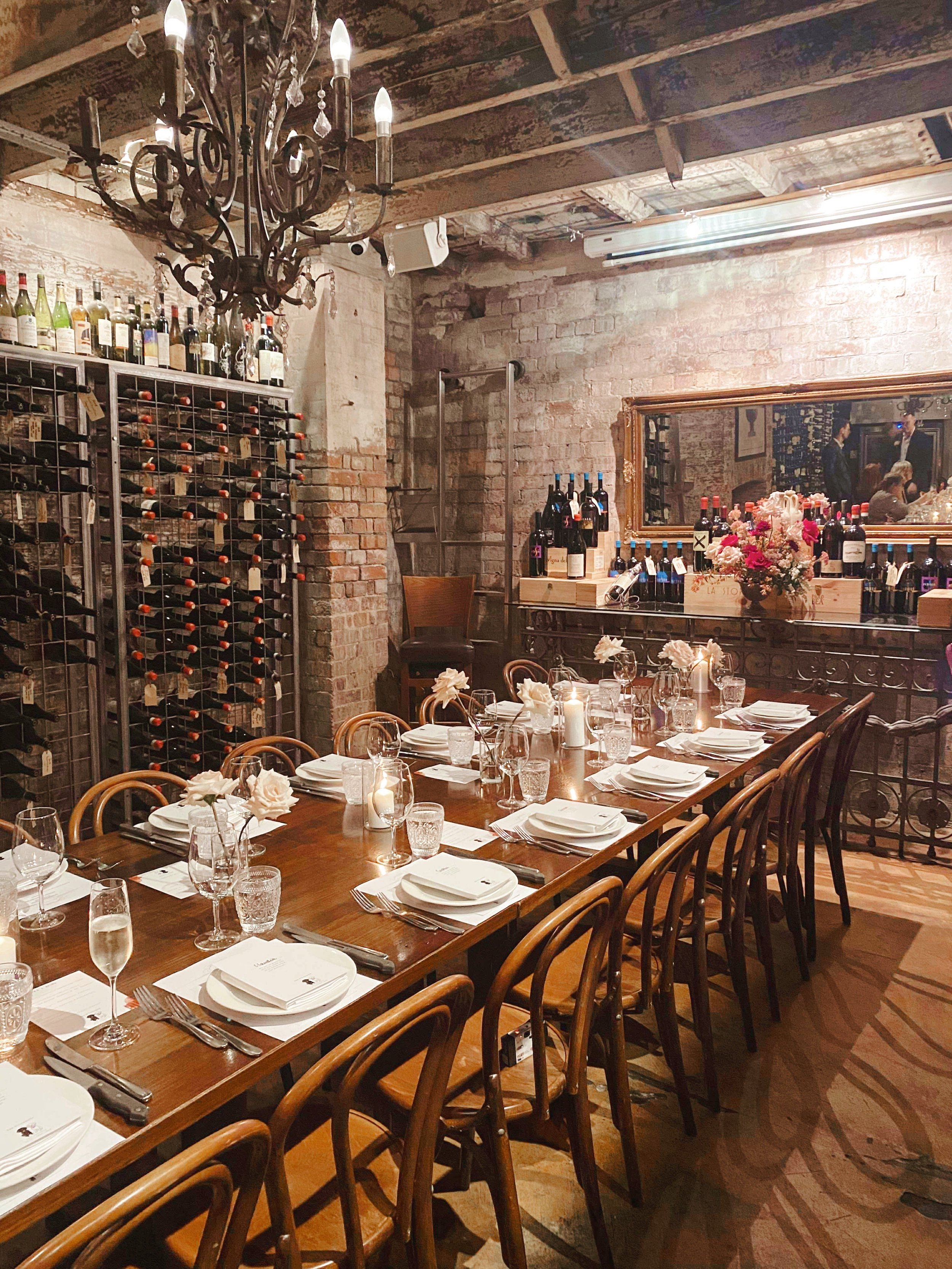 A cozy restaurant with a long wooden dining table set for a meal, with wine glasses, white napkins, and candles, surrounded by wooden chairs. There is a large mirror and a wine rack against brick walls, with wine bottles displayed on shelves and in r