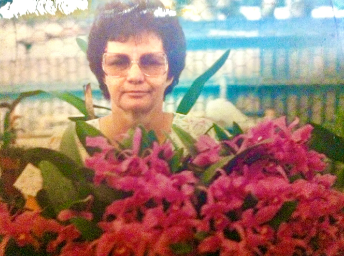 A woman with dark hair, sunglasses, and a light-colored top, standing behind a large bouquet of pink flowers with green leaves, outdoors with a fence and trees in the background.