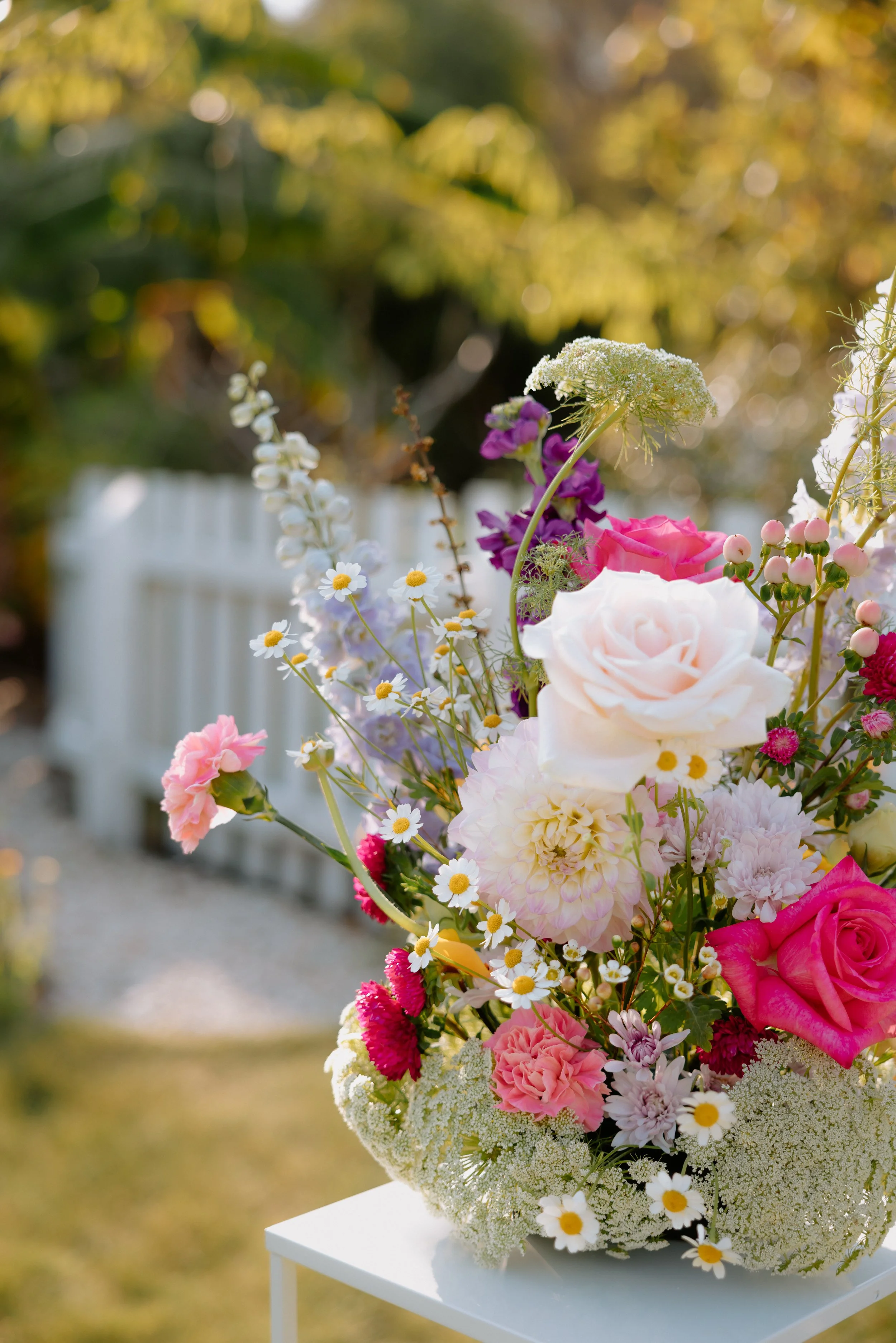 A colorful bouquet of various flowers, including roses, daisies, and hydrangeas, placed on a white table outdoors with a blurred background of greenery and a white picket fence.