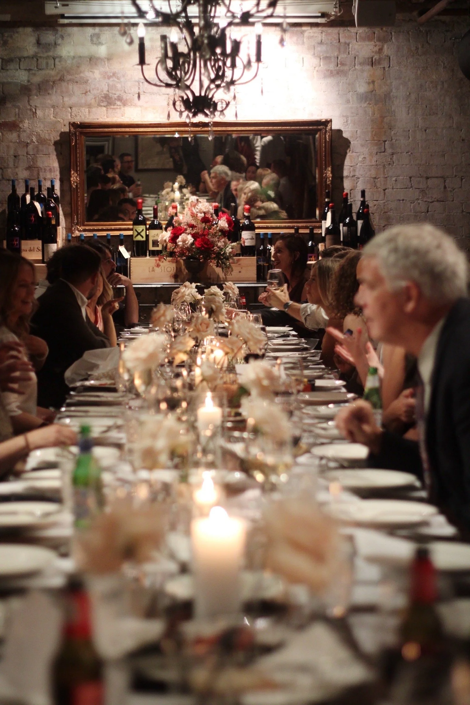People dining at Enoteca Brisbane at a long, elegant dinner table decorated with white flowers and candles, in a restaurant with a brick wall and a large mirror, bottles of wine visible on a sideboard.