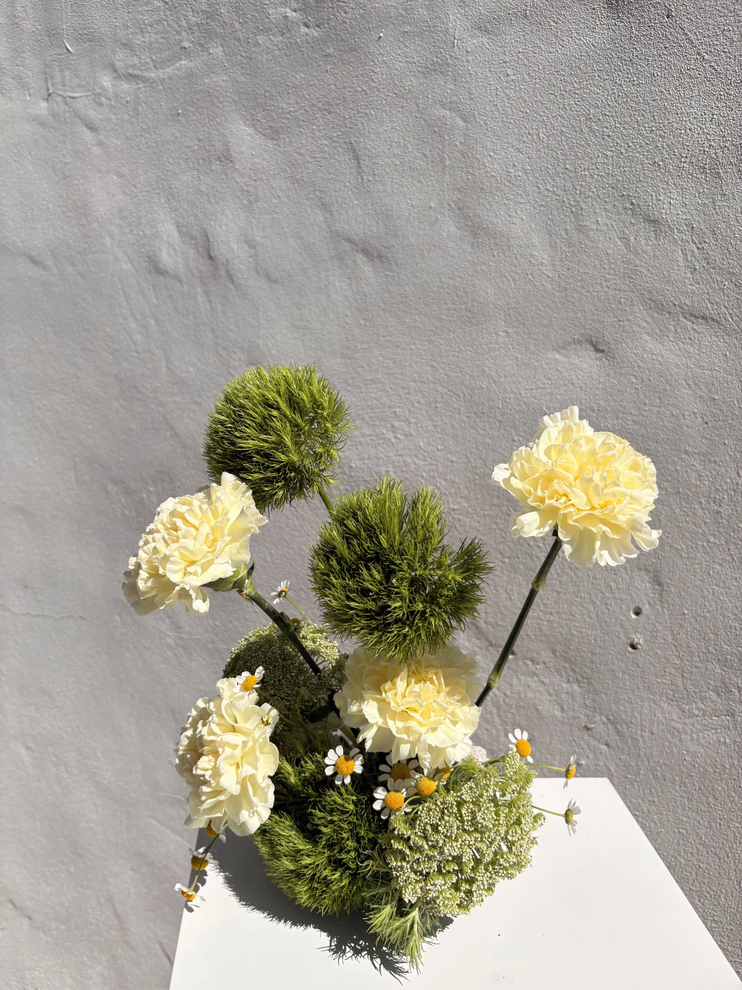 Arrangement of white carnations, green pom-pom flowers, and small white daisies in a white vase on a white surface against a textured white wall.