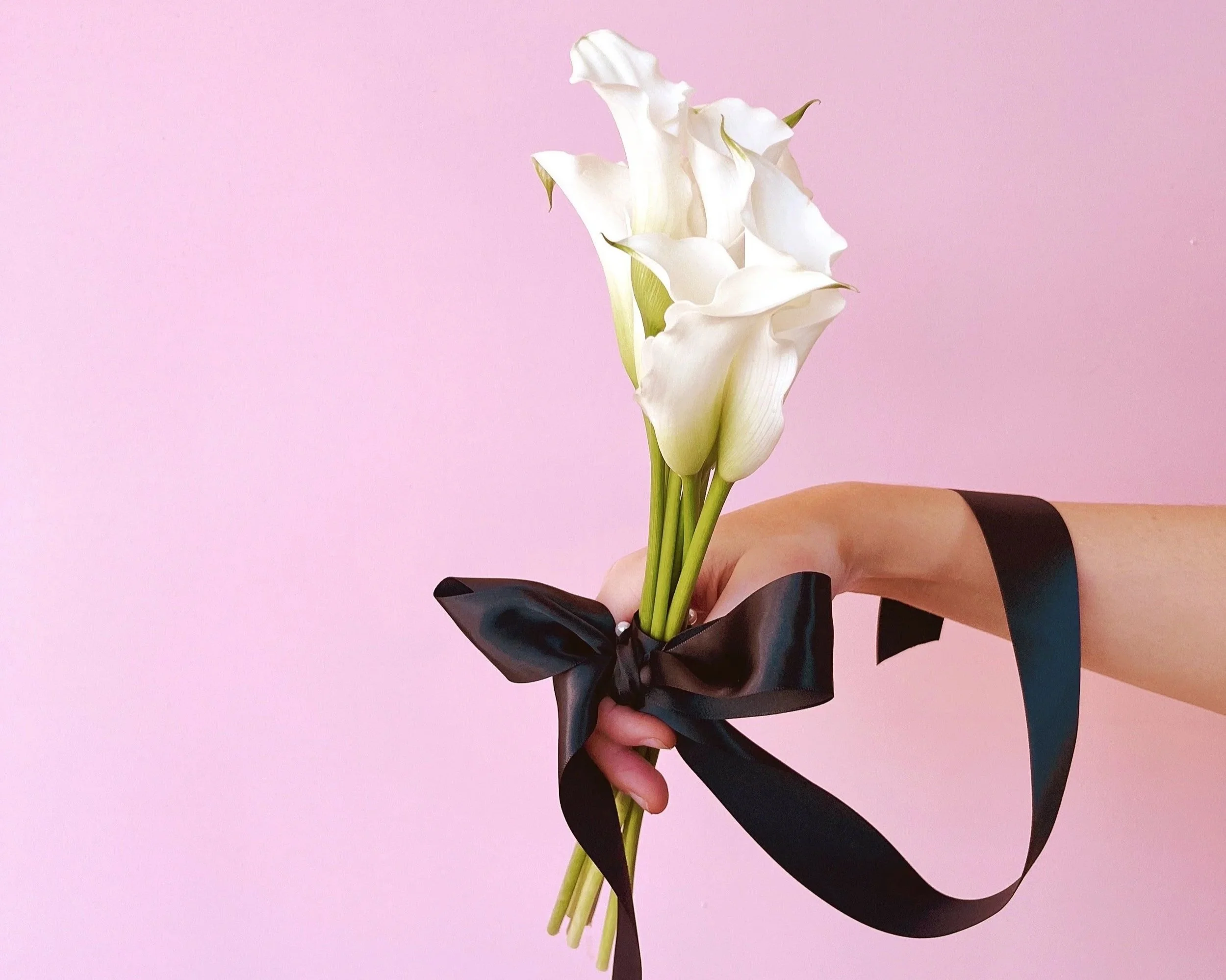 Brisbane Florist, From June, with a hand holding white calla lilies tied with a black satin ribbon against a pink background.