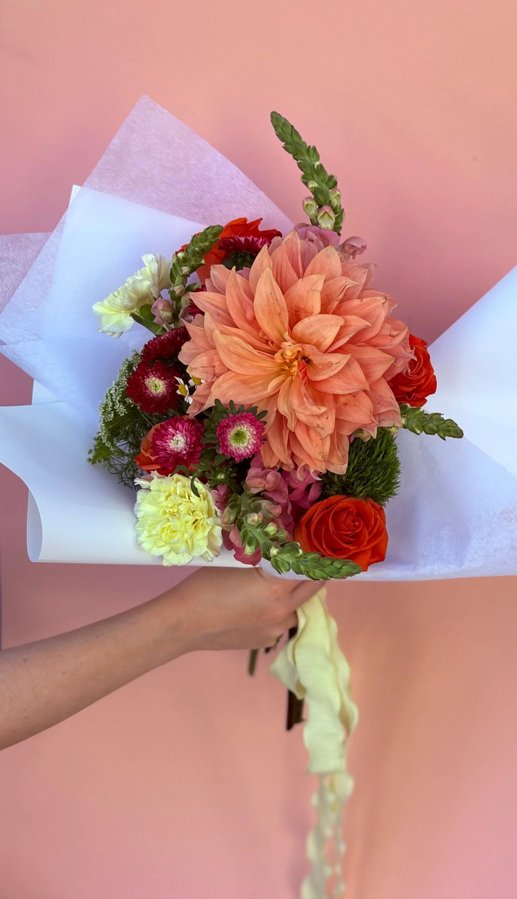 Colorful mixed flower bouquet including peach dahlia, orange roses, pink and red mums, green foliage, wrapped in white paper, held by a person against a pink background.
