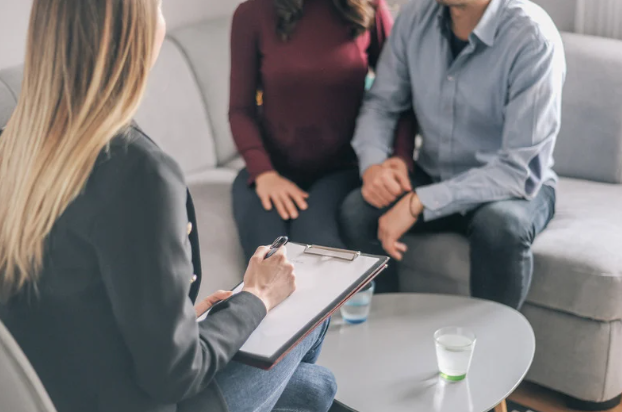 A woman in a gray blazer sits with a clipboard, taking notes during a therapy or counseling session with a couple sitting on a couch.
