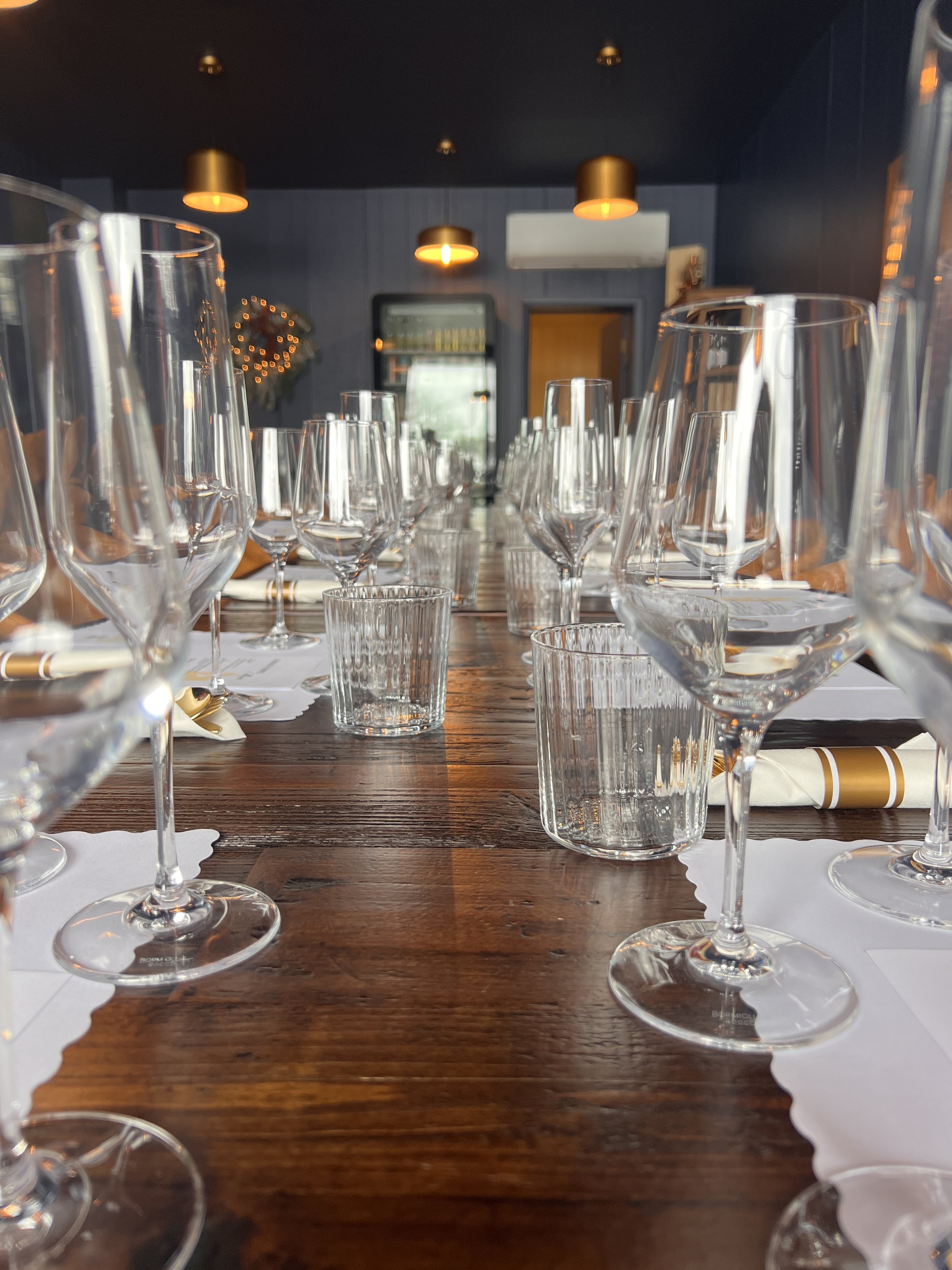 Table set for a formal dinner with glasses, napkins, and utensils in a restaurant with dark walls and warm lighting.