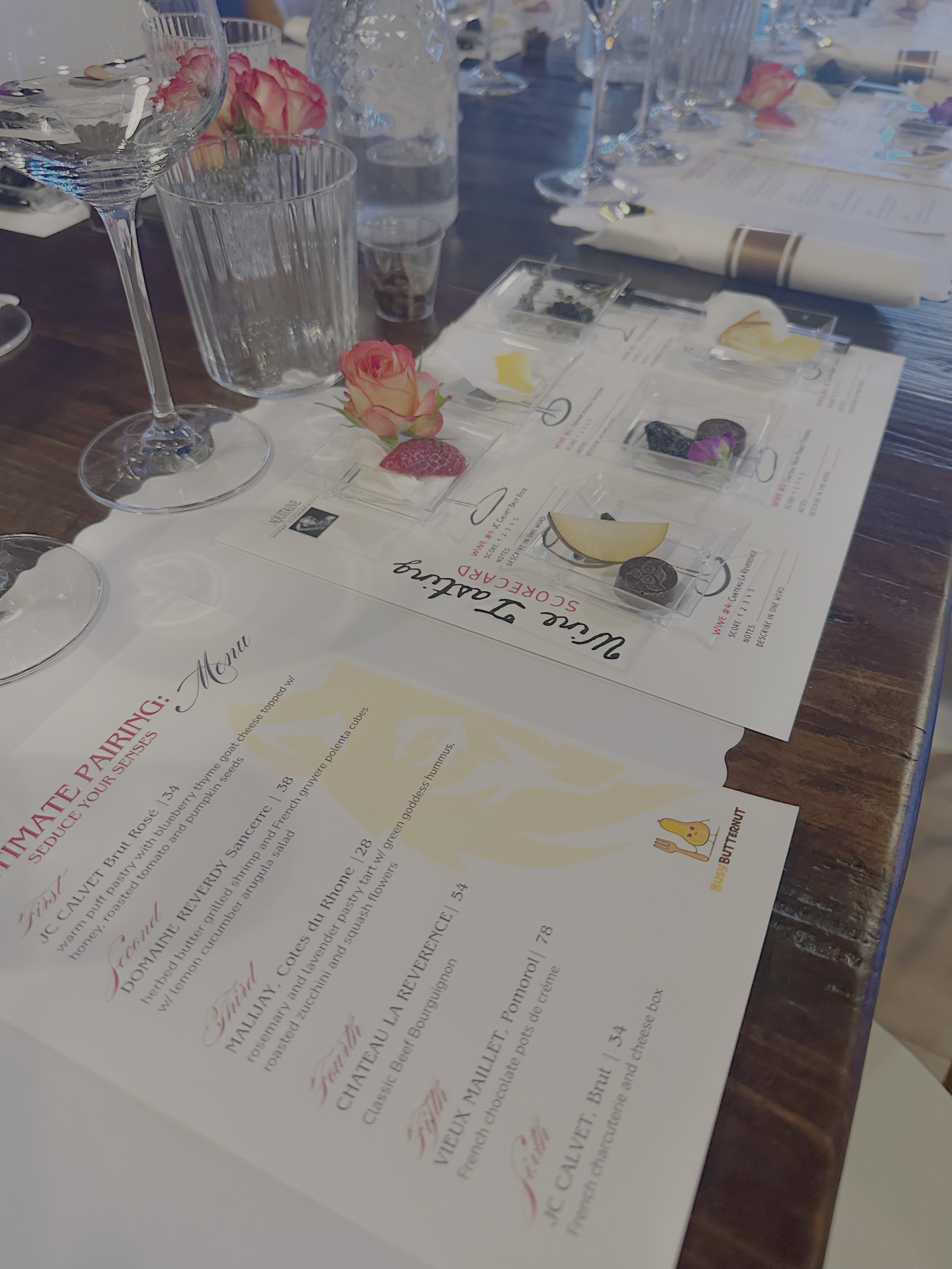 A table set with glasses, water bottles, pink roses, and a menu featuring a wine and food pairing.