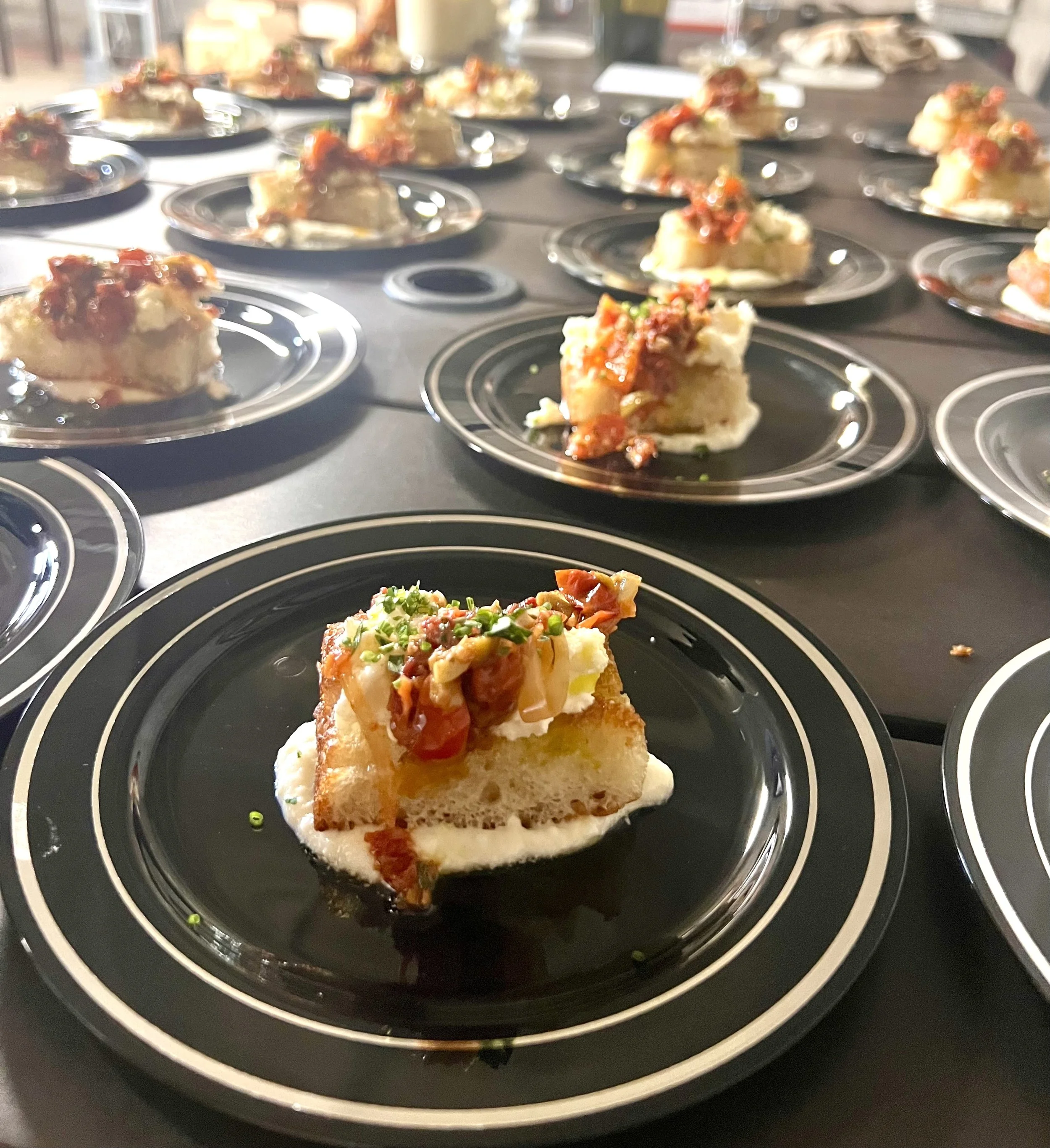 A close-up of food tapas on black and white serving plates, featuring small sandwiches topped with shredded lettuce and cherry tomatoes, on a stove top with multiple burners.