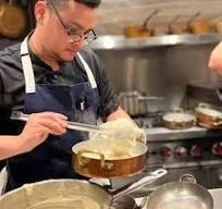 A chef in a professional kitchen is lifting a dish from a frying pan using a spatula.
