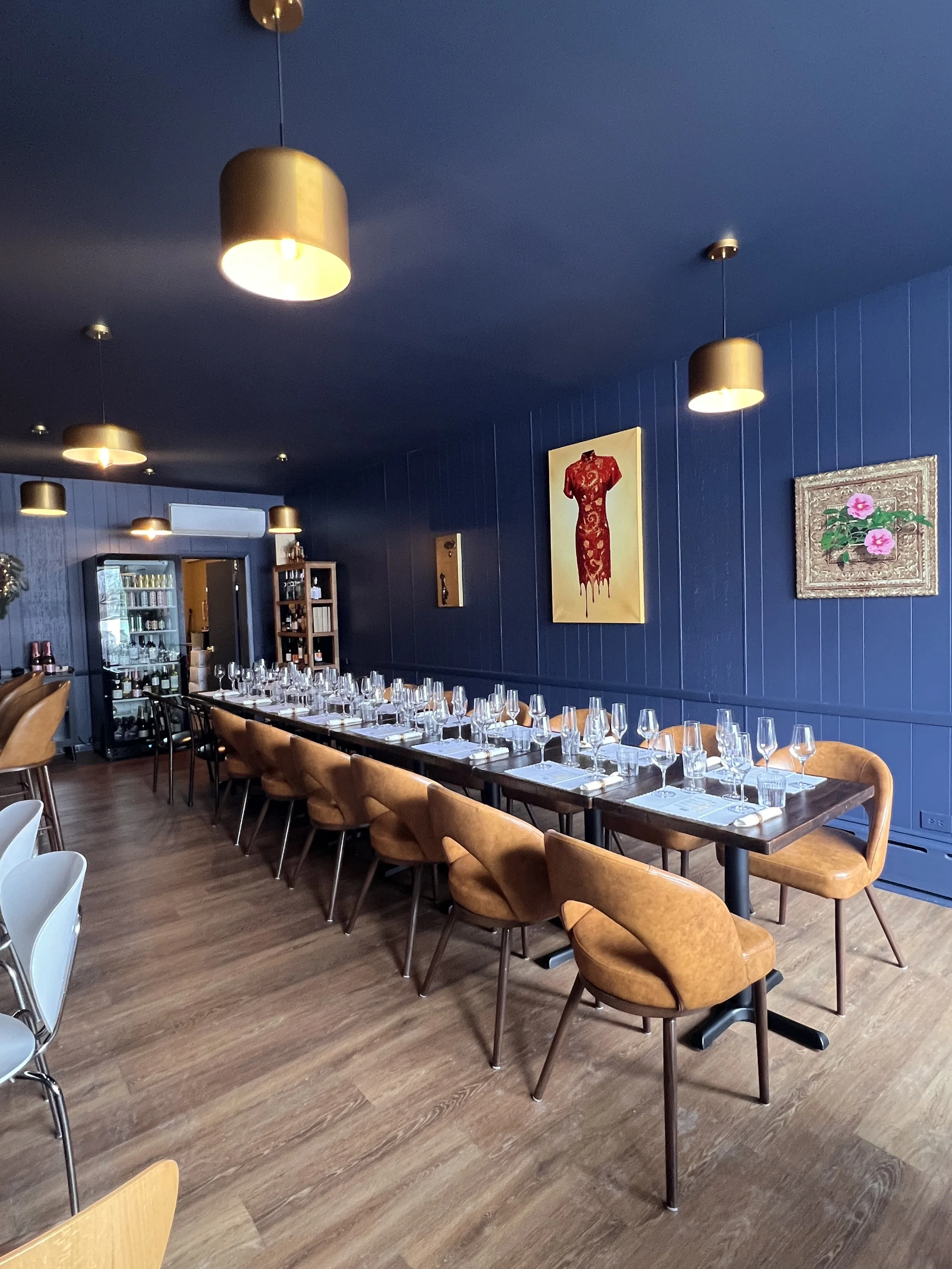A long dining table with settings for many guests, surrounded by tan and black chairs, in a restaurant with dark blue walls decorated with paintings and art.