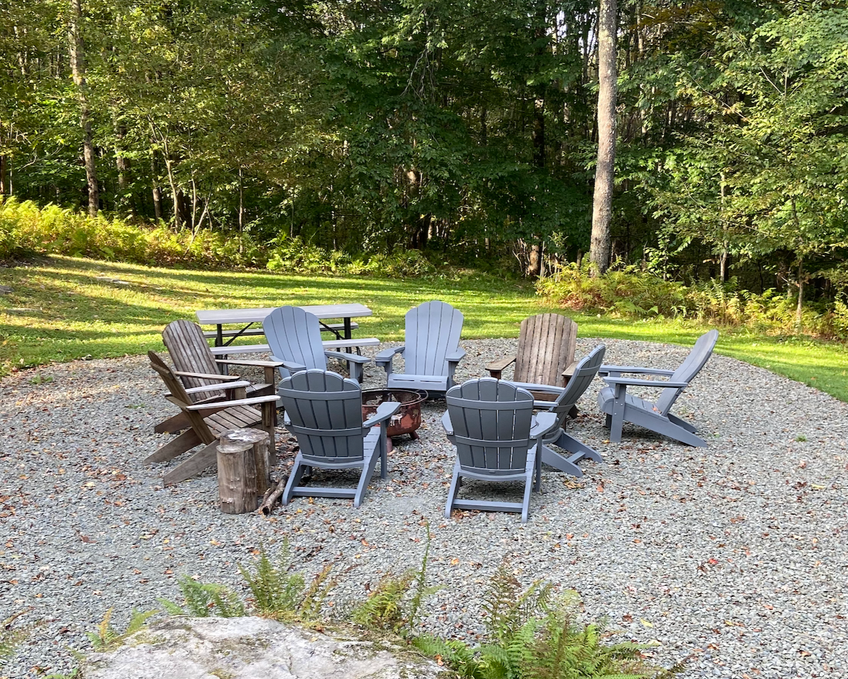 Eight adirondack chairs circle a firepit on gravel in the woods inviting IFS group consultation.