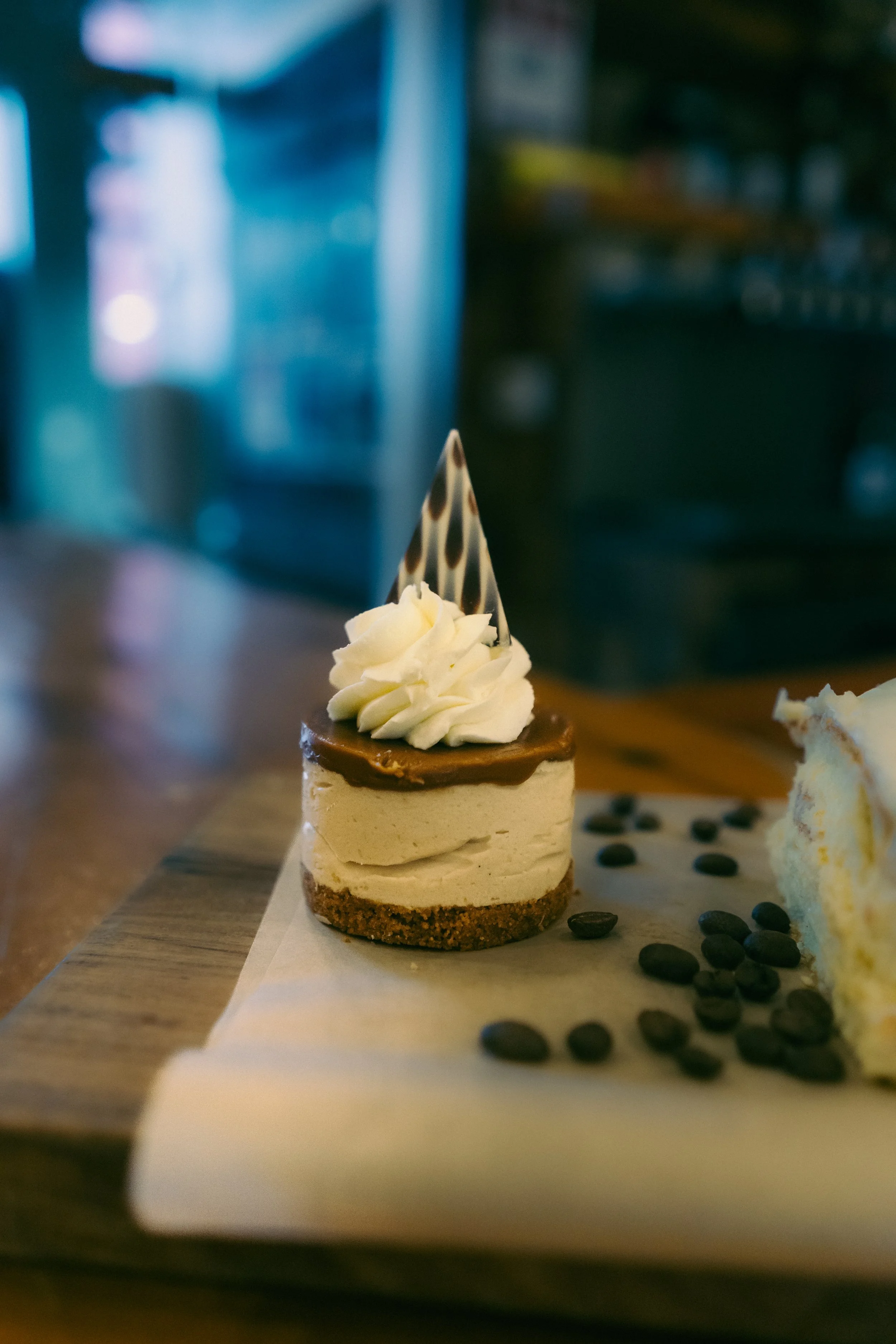 Small, layered cheesecake with whipped cream and a chocolate and white decorative triangle on top, served on a wooden platter with scattered coffee beans.