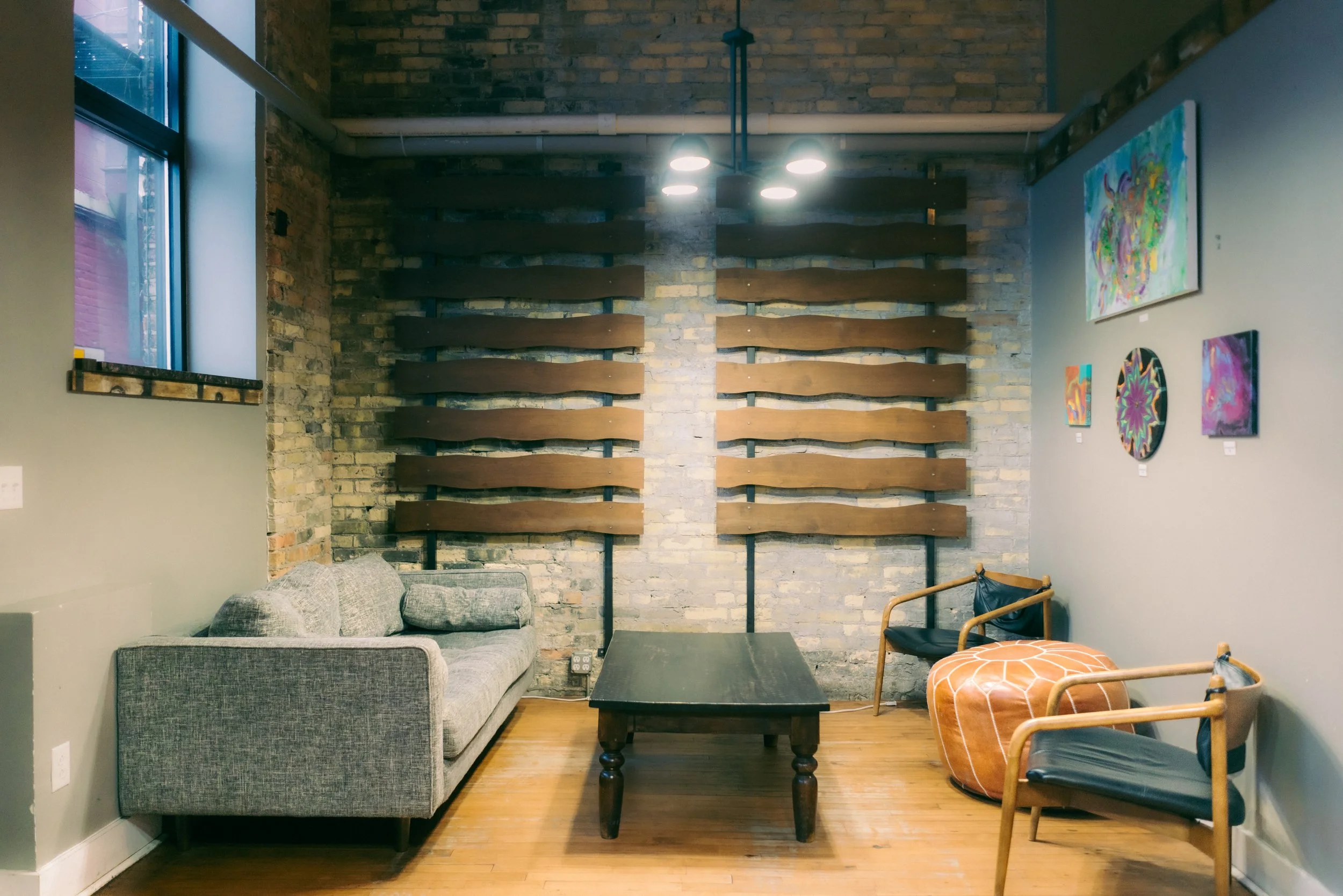 Art gallery with a brick wall display of wooden slats behind a gray sofa, two chairs, a coffee table, and colorful artwork on the walls.