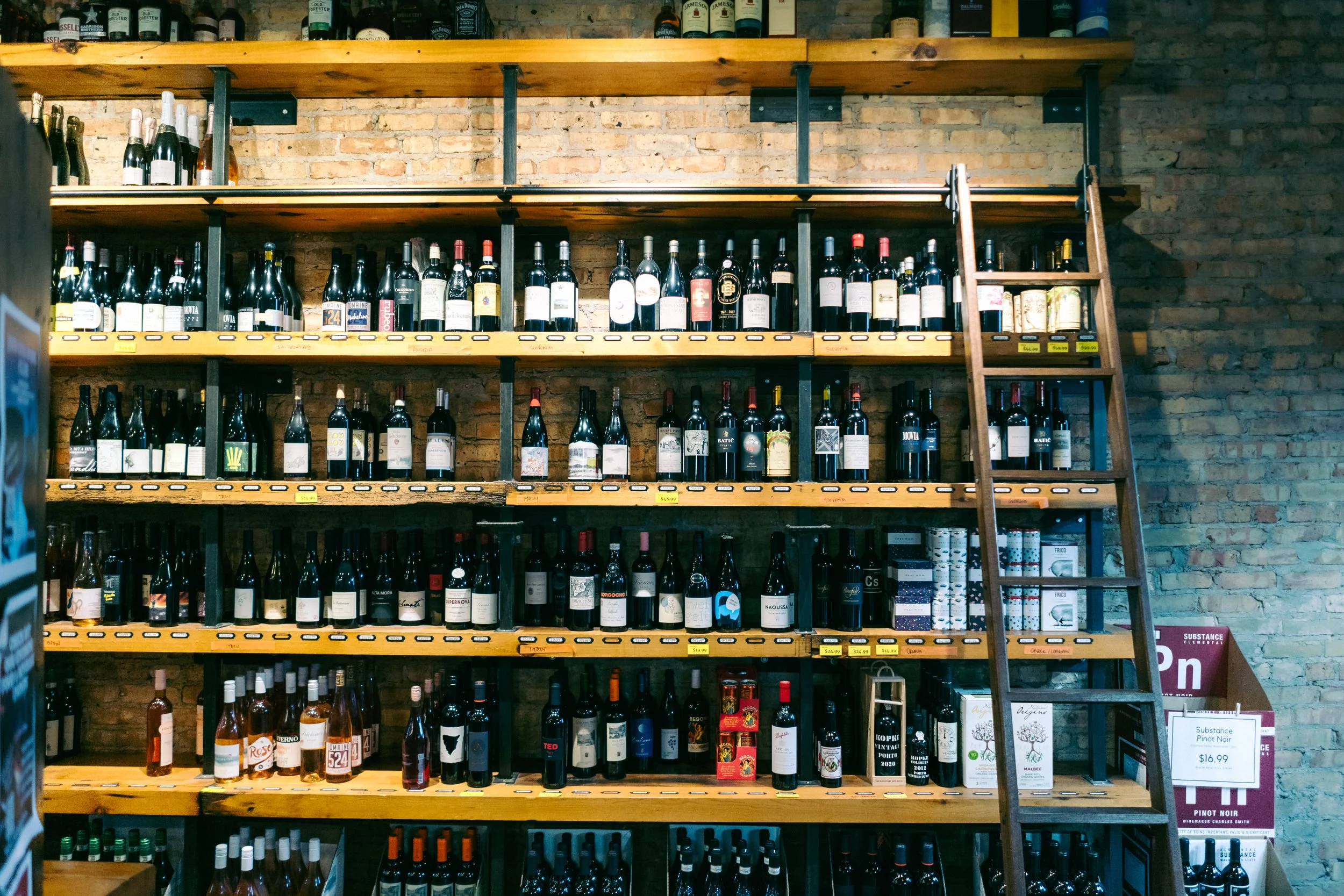 Shelves stocked with various bottles of wine and alcohol against a brick wall, with a wooden ladder on the right side and a sign indicating a Pinot Noir for $16.99.