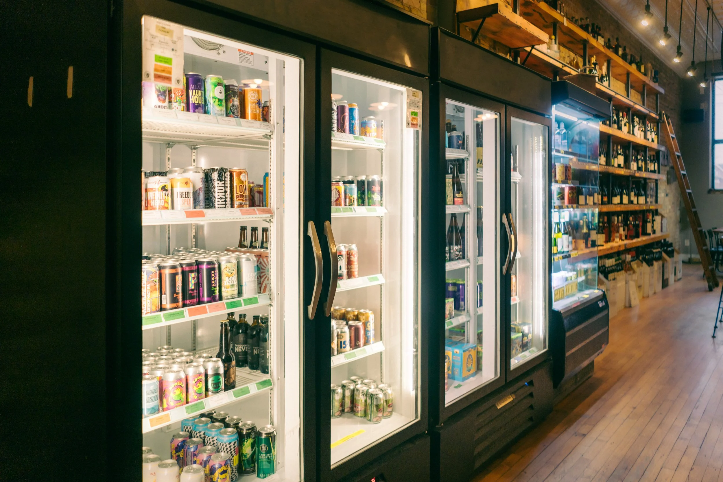 Refrigerators filled with a variety of canned and bottled beverages, located in a modern store or cafe with wooden floors and shelves of wine bottles on the wall.