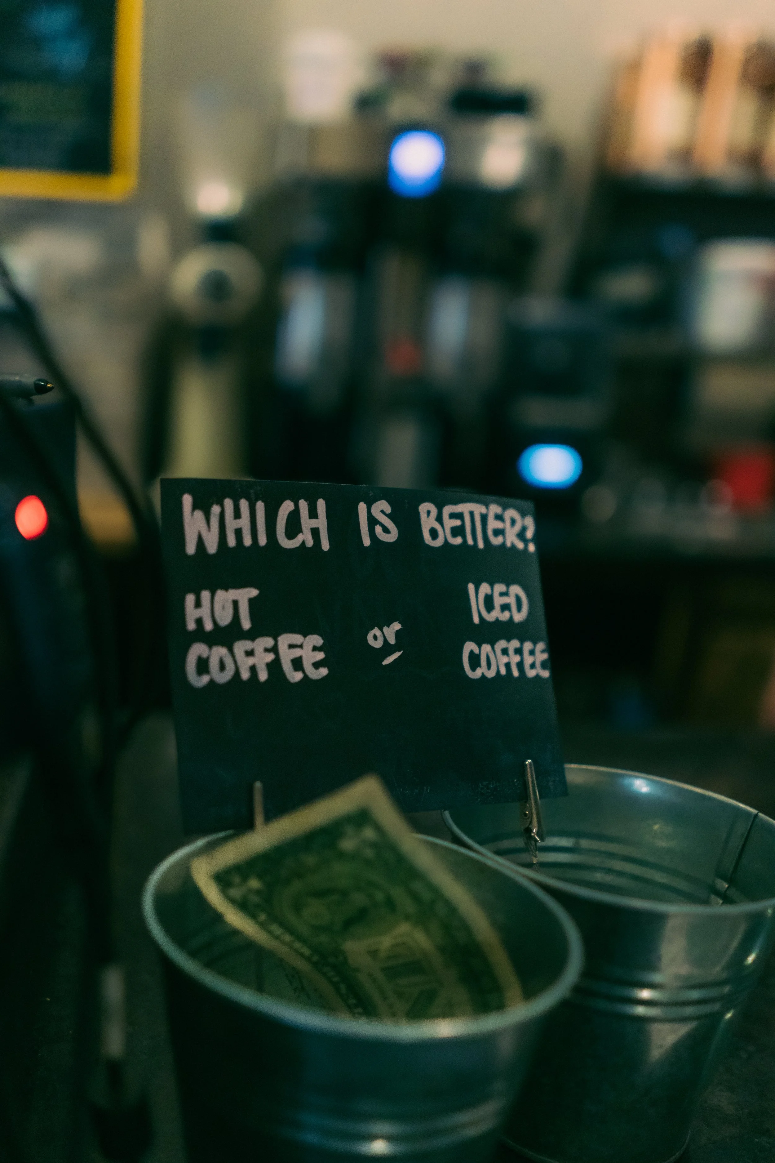 A sign asking 'Which is better? Hot coffee or iced coffee?' placed in metal buckets containing dollar bills, with a blurred coffee shop background.