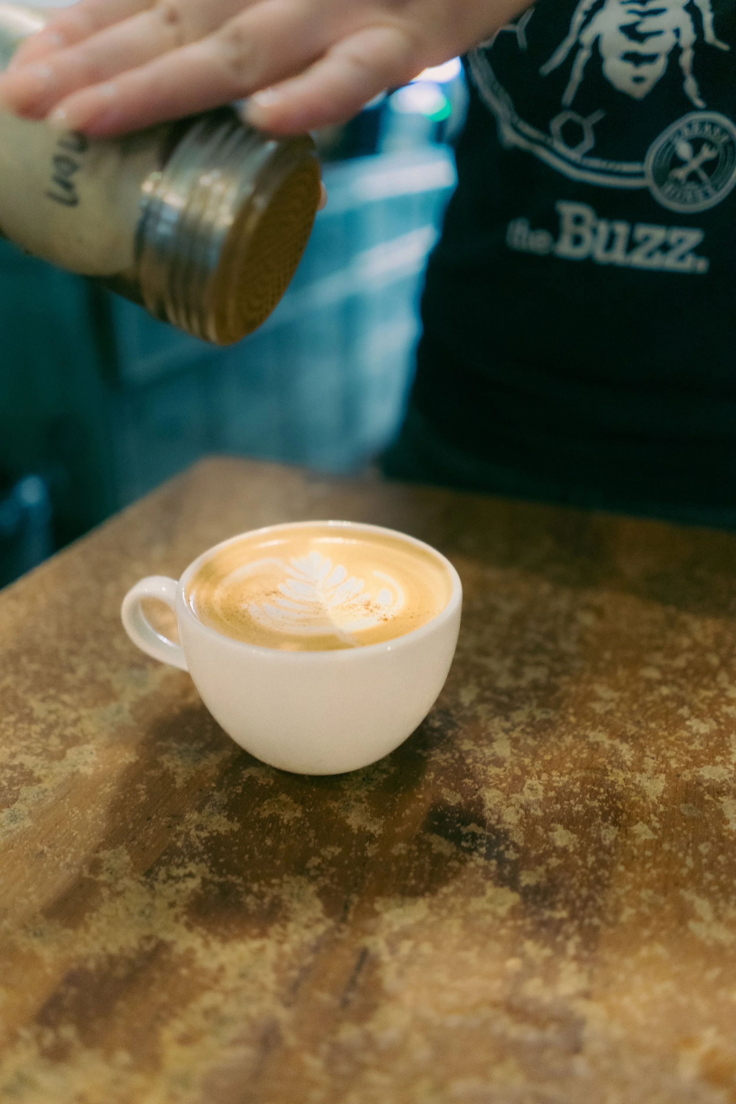 A person pouring caramel syrup onto a latte in a white ceramic cup on a wooden table.