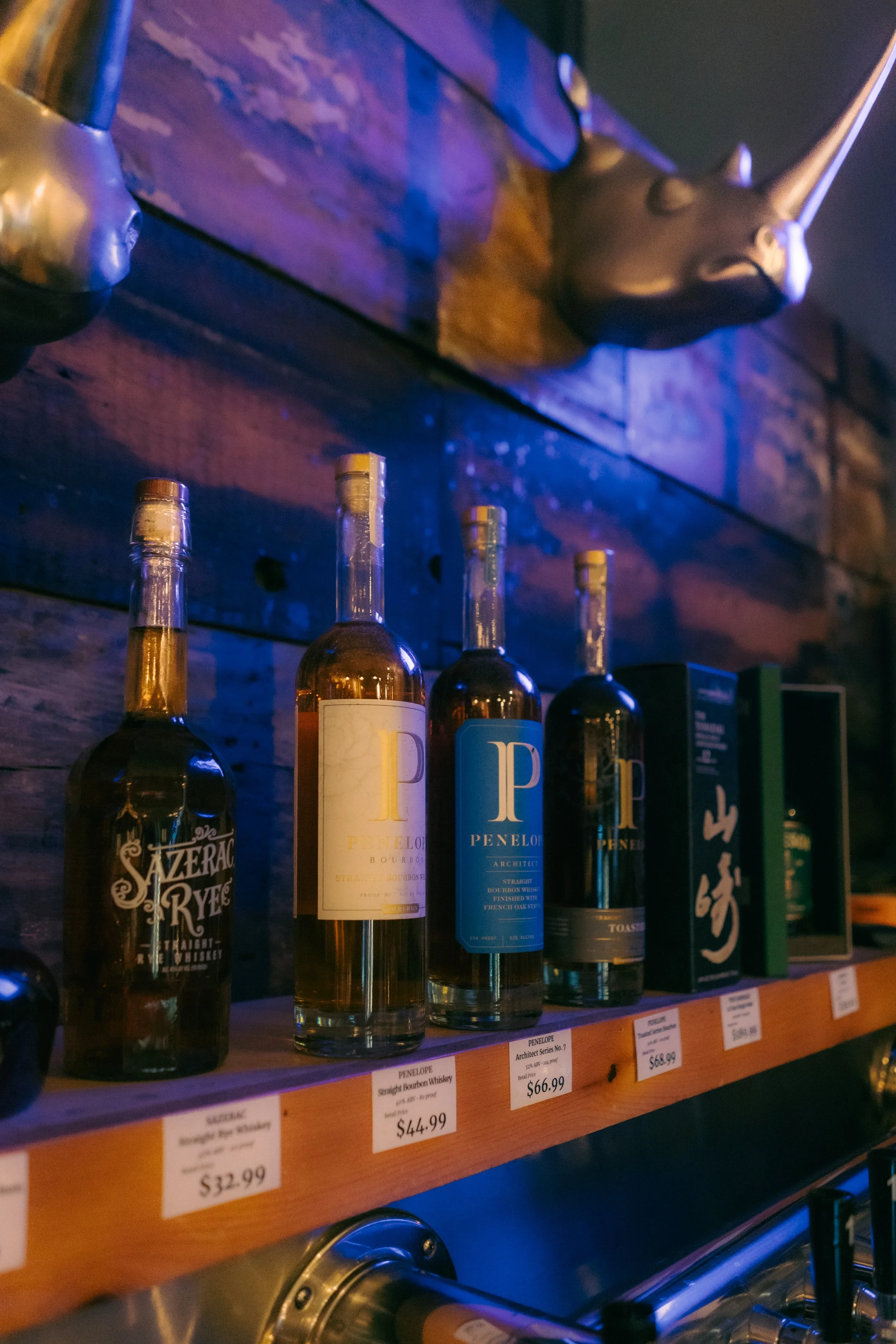 A row of liquor bottles on a wooden shelf, including brands like Penelope bourbon andazerac rye, with price tags below. A unicorn head sculpture is mounted on a rustic wooden wall above the bottles, illuminated by dim lighting.
