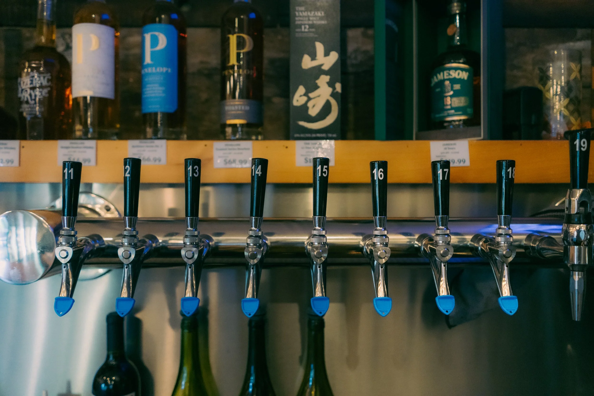 Seven beer taps with black handles numbered 11 to 17, mounted on a silver tap wheel, with bottles hanging beneath. In the background, various bottles of liquor are displayed on a wooden shelf.