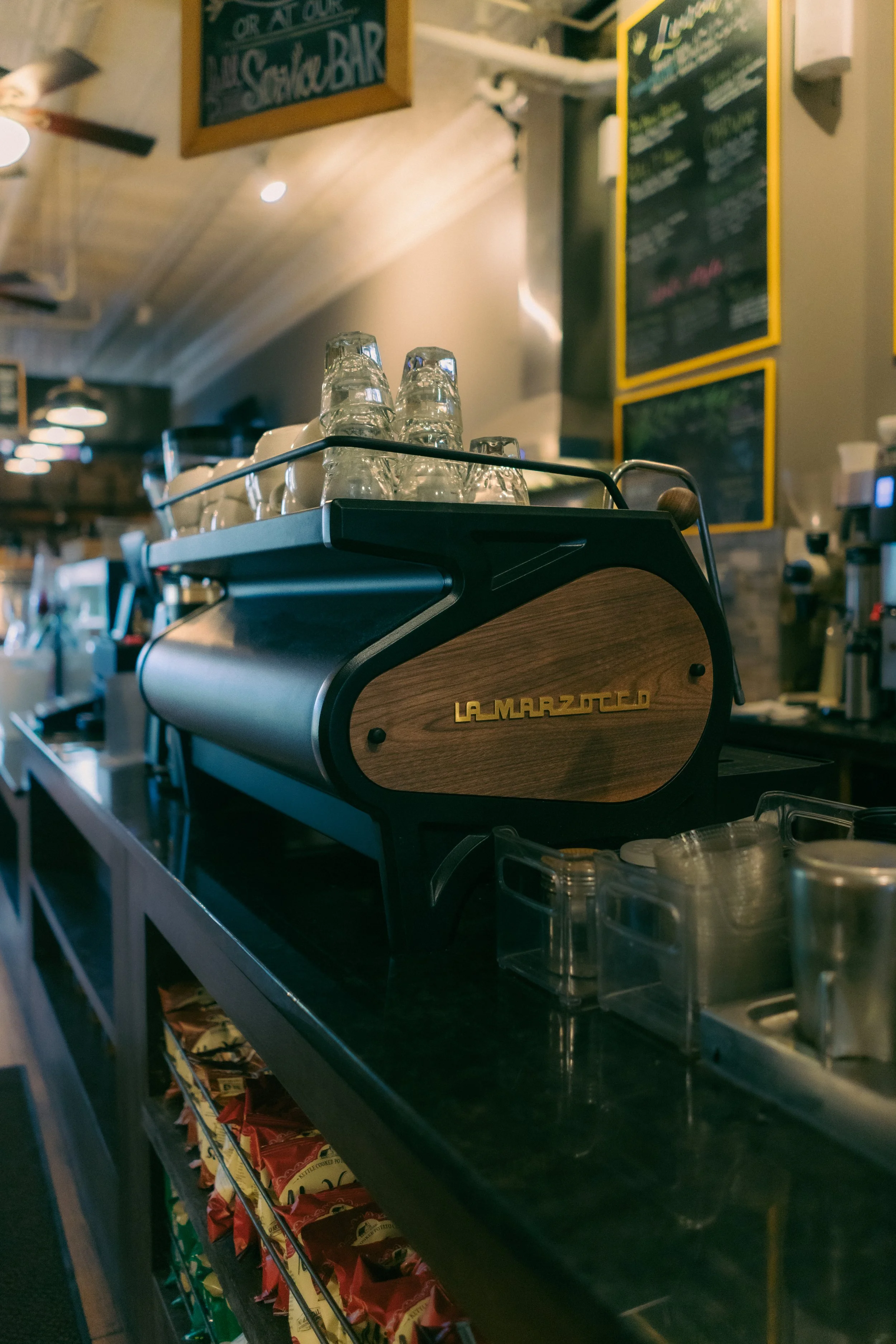 Coffee shop countertop with a La Marzocco espresso machine and glass cups on top, menu chalkboards in the background.