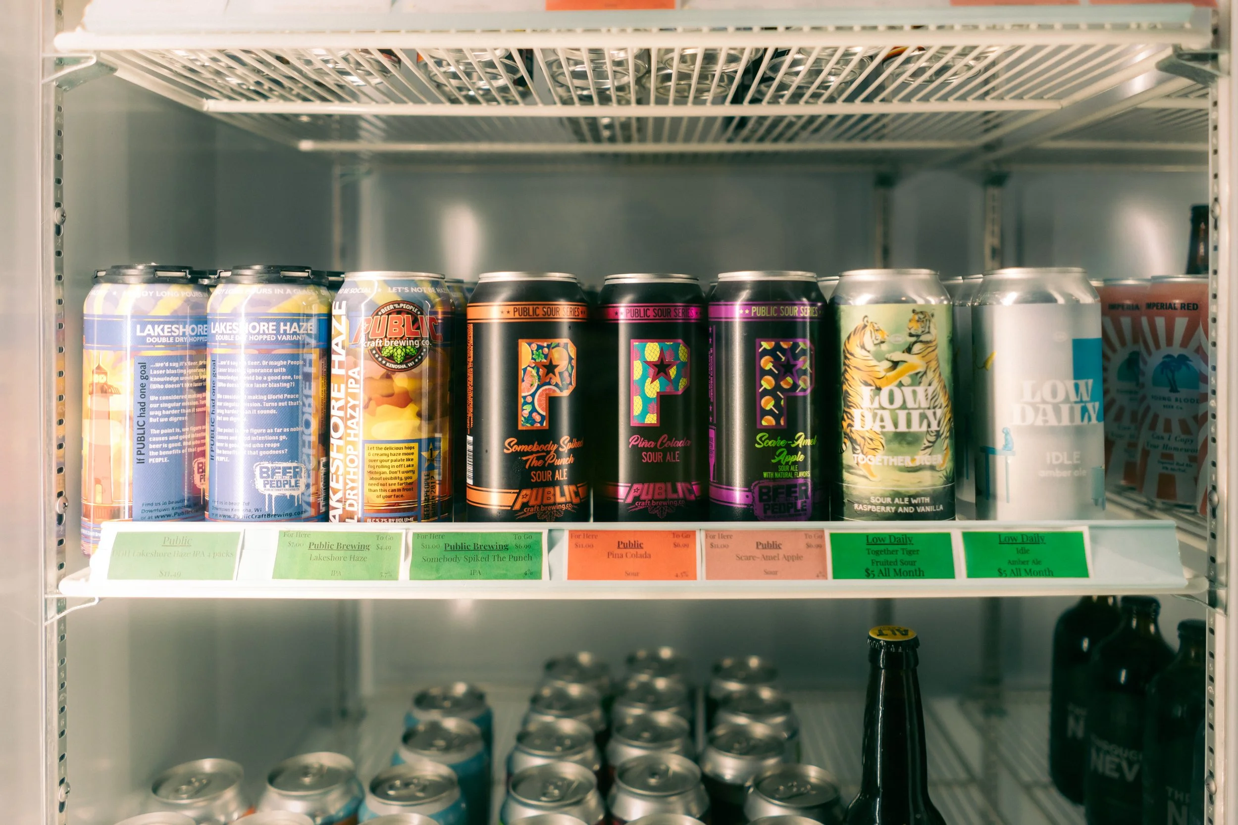 A refrigerator shelf with various cans of craft beer, including Lakeshore Haze, Public, and Low Daily.