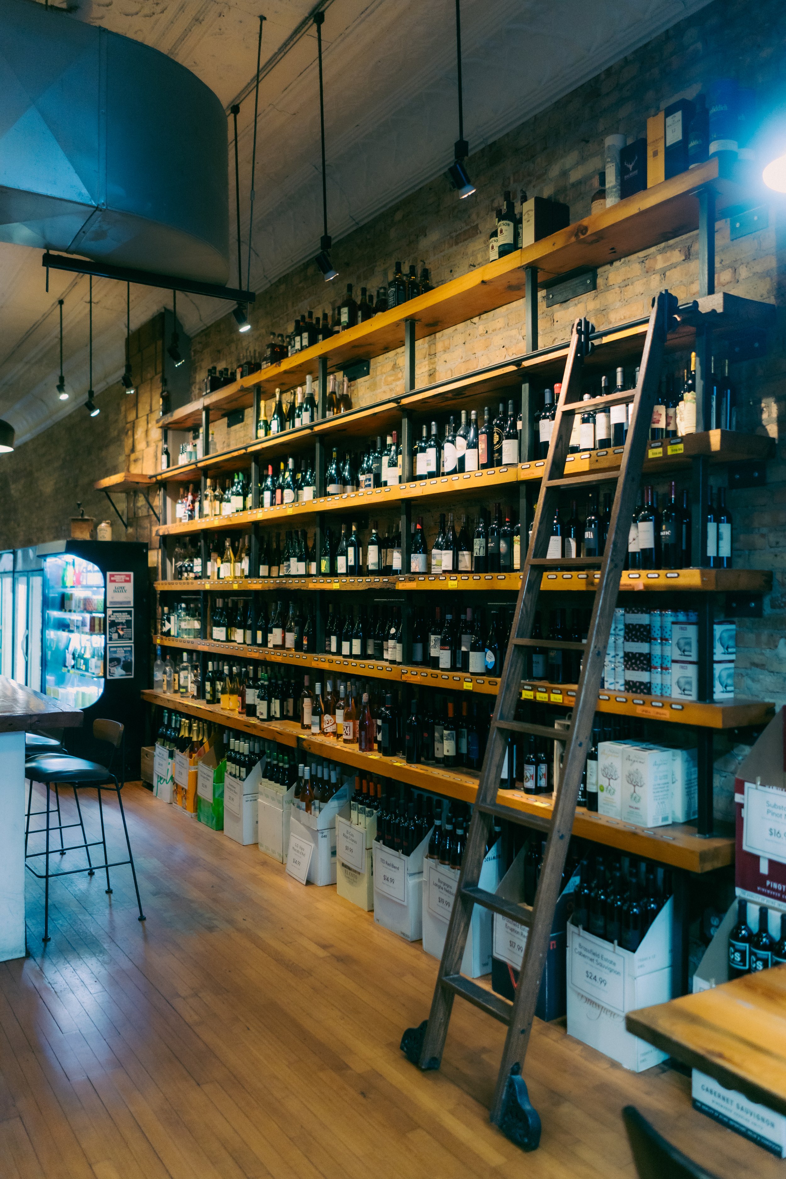 Interior of a store with wooden shelves stocked with bottles of wine and liquor, a ladder leaning against the shelves, and a vending machine on the left side.