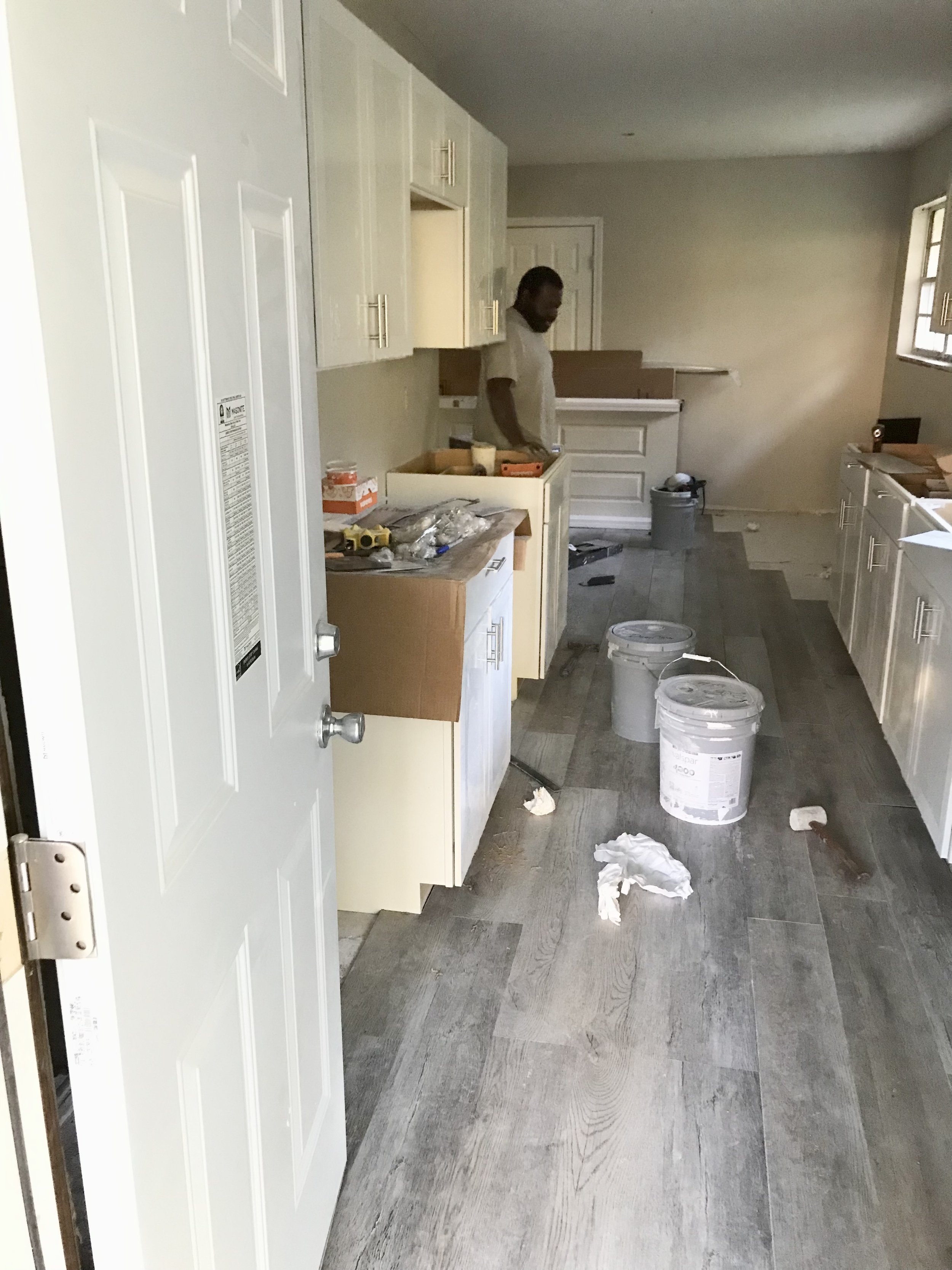 A man standing in a kitchen under construction or renovation, with white cabinets, a window on the right, and construction supplies and debris on the floor.