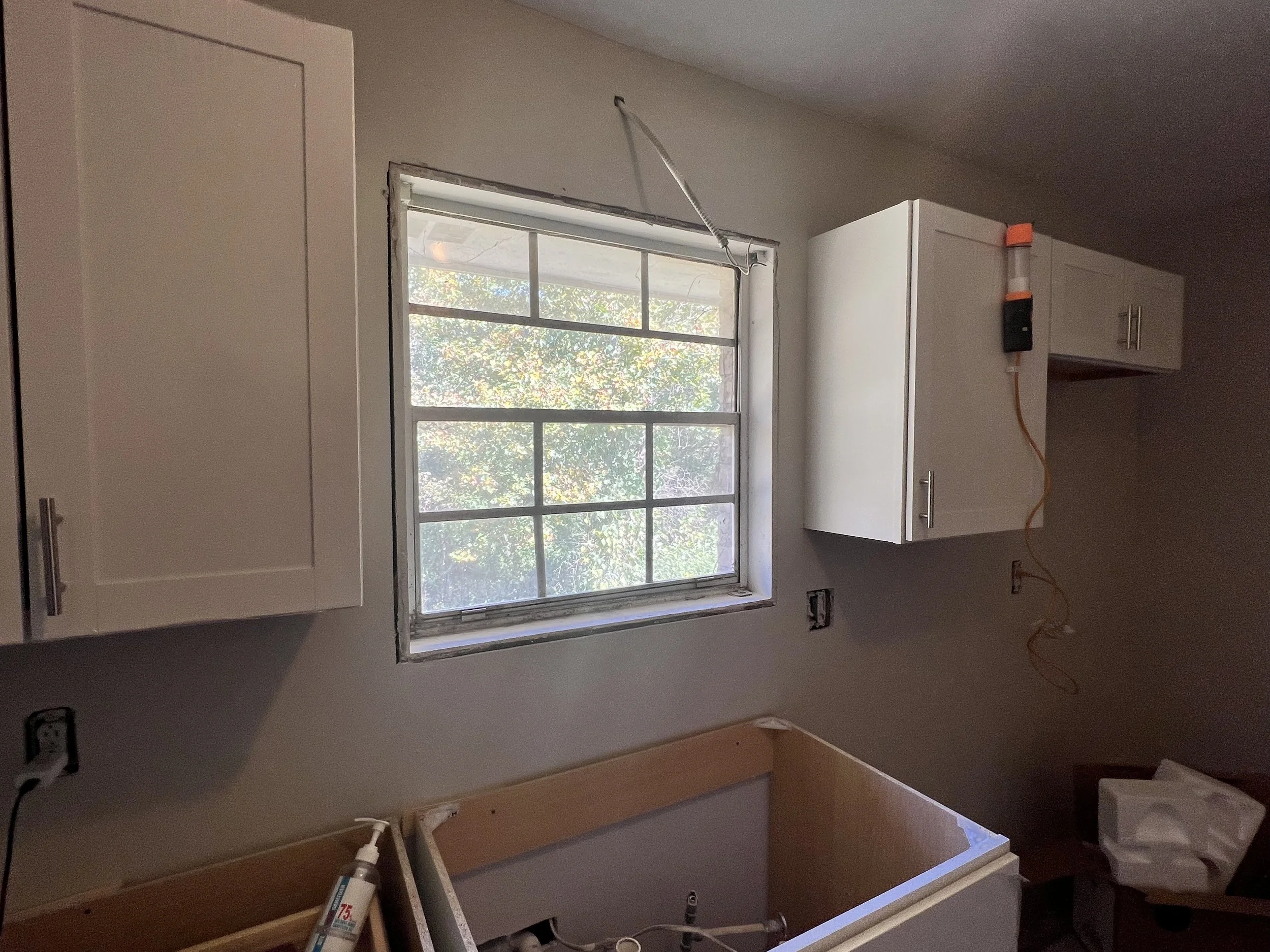 Kitchen renovation in progress showing a window with partially installed cabinets on either side, construction tools, and an empty space where a sink or appliance will be installed.