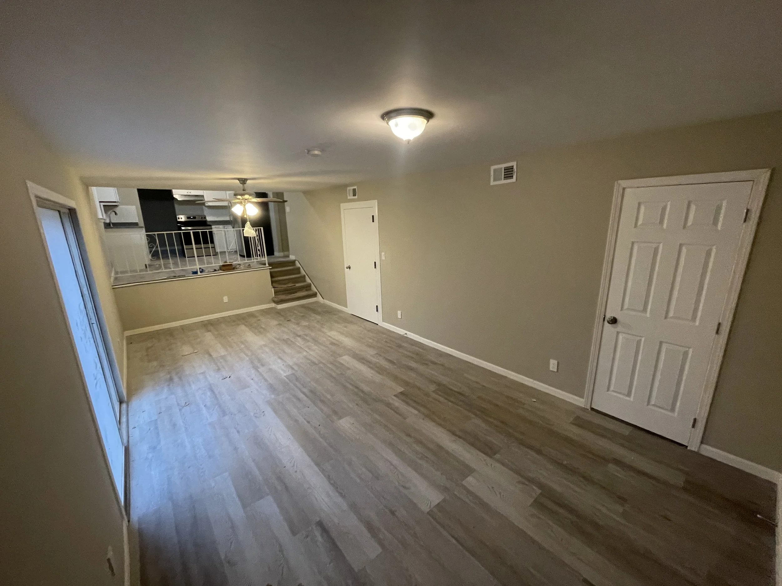 Empty living room with beige walls, wood laminate flooring, white baseboards, a ceiling light, and sliding glass door leading outside. A staircase with carpeted steps leads into the room, and kitchen area is visible in the background with appliances,