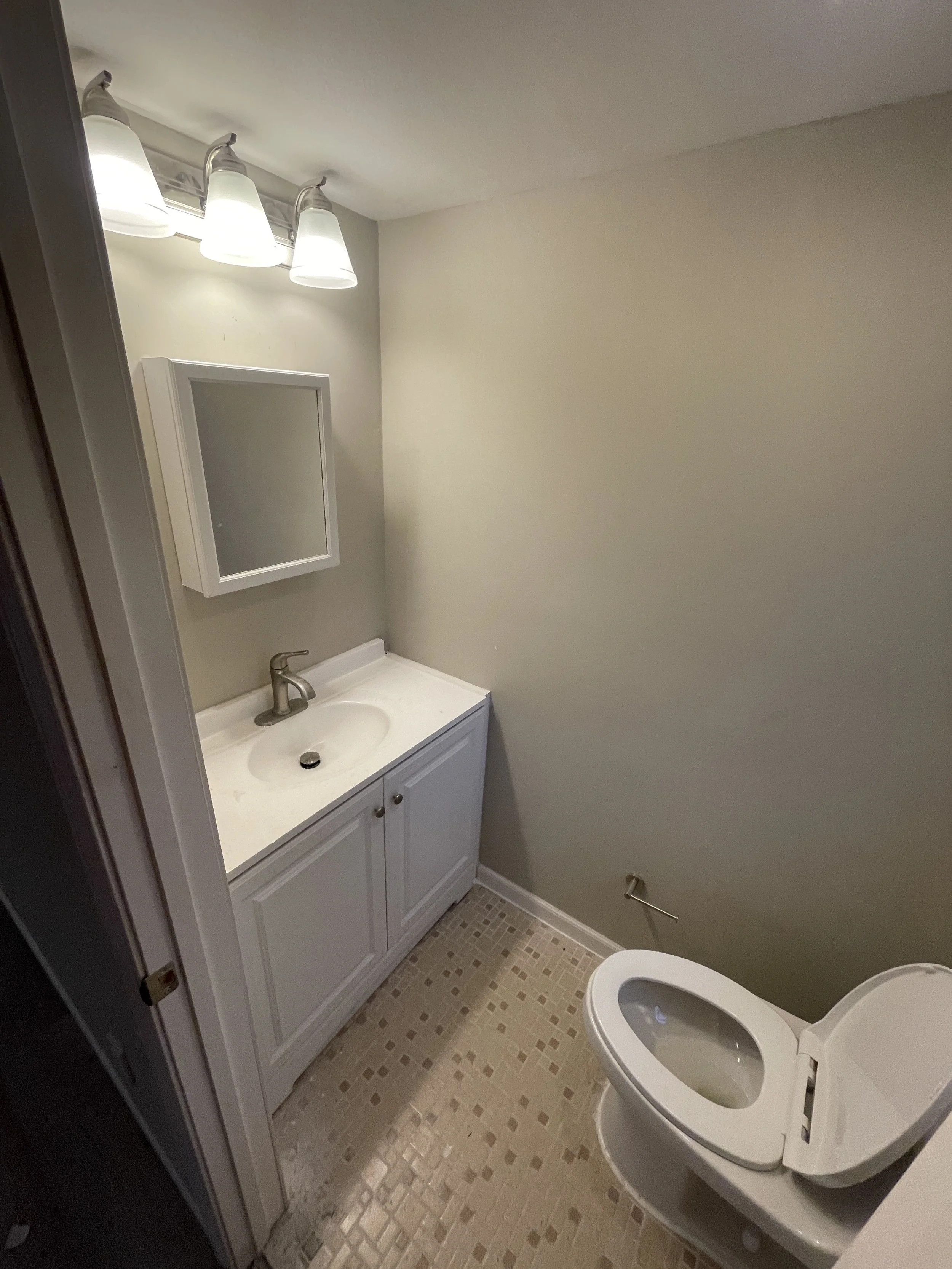 Small bathroom with a white vanity sink, a mirror above the sink, a light fixture with four lights above the mirror, and a toilet to the right. The floor has beige mosaic tiles.