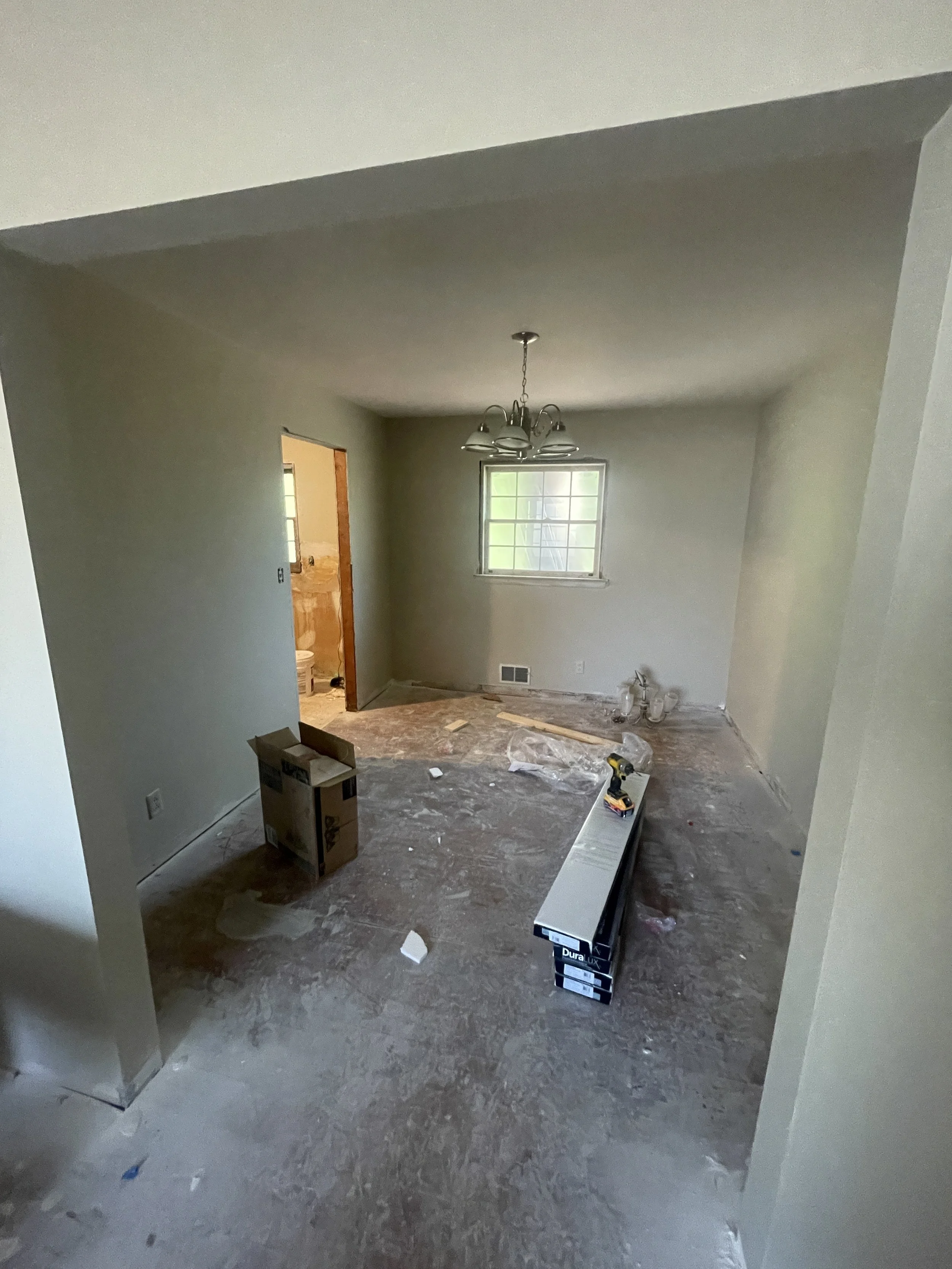 Empty living room with a window, chandelier, and ongoing renovation including tools, a cardboard box, and construction materials on the floor.