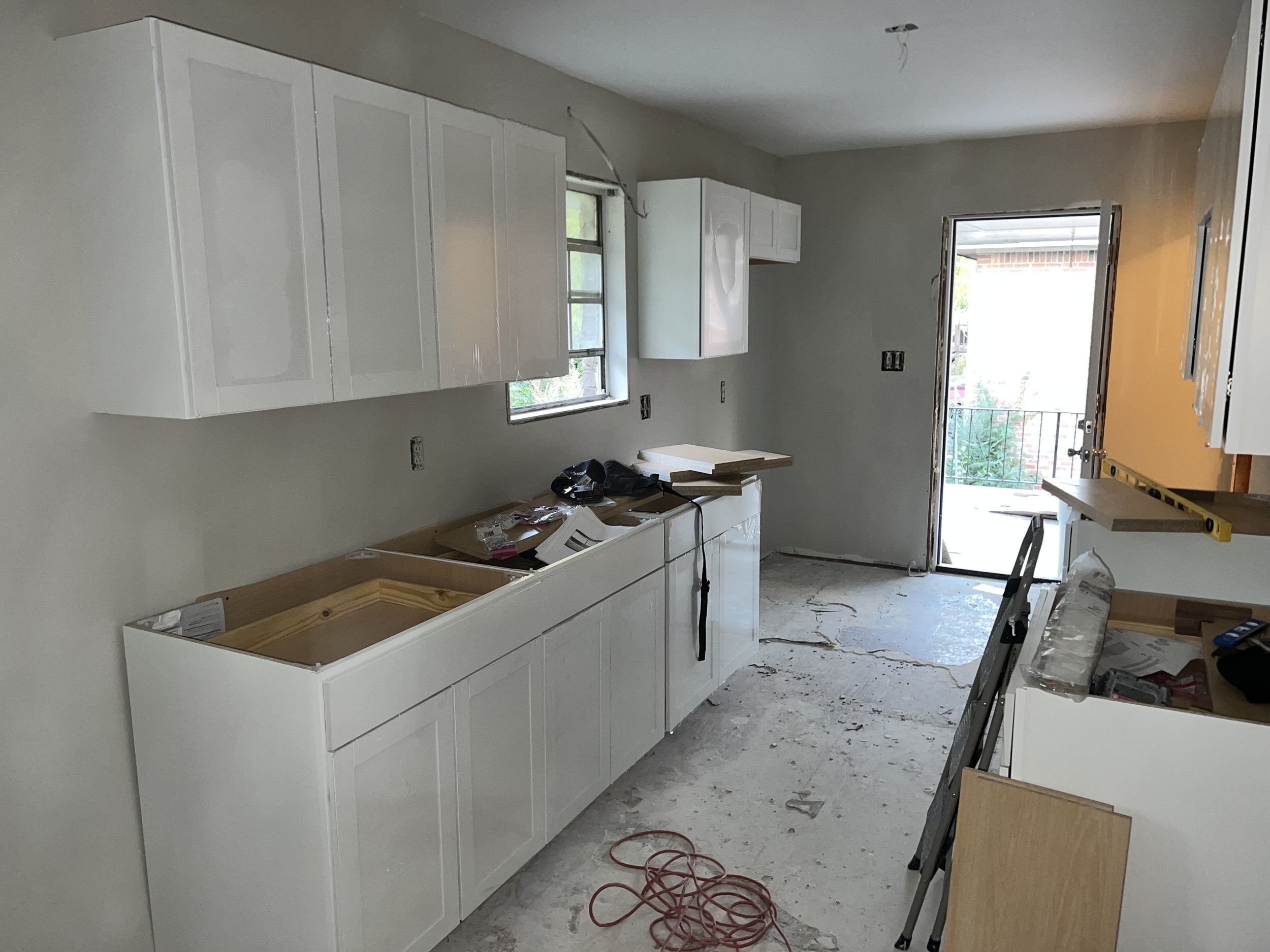 Kitchen under construction with white cabinets, a window, an open door leading to a balcony, construction tools, and unfinished flooring.