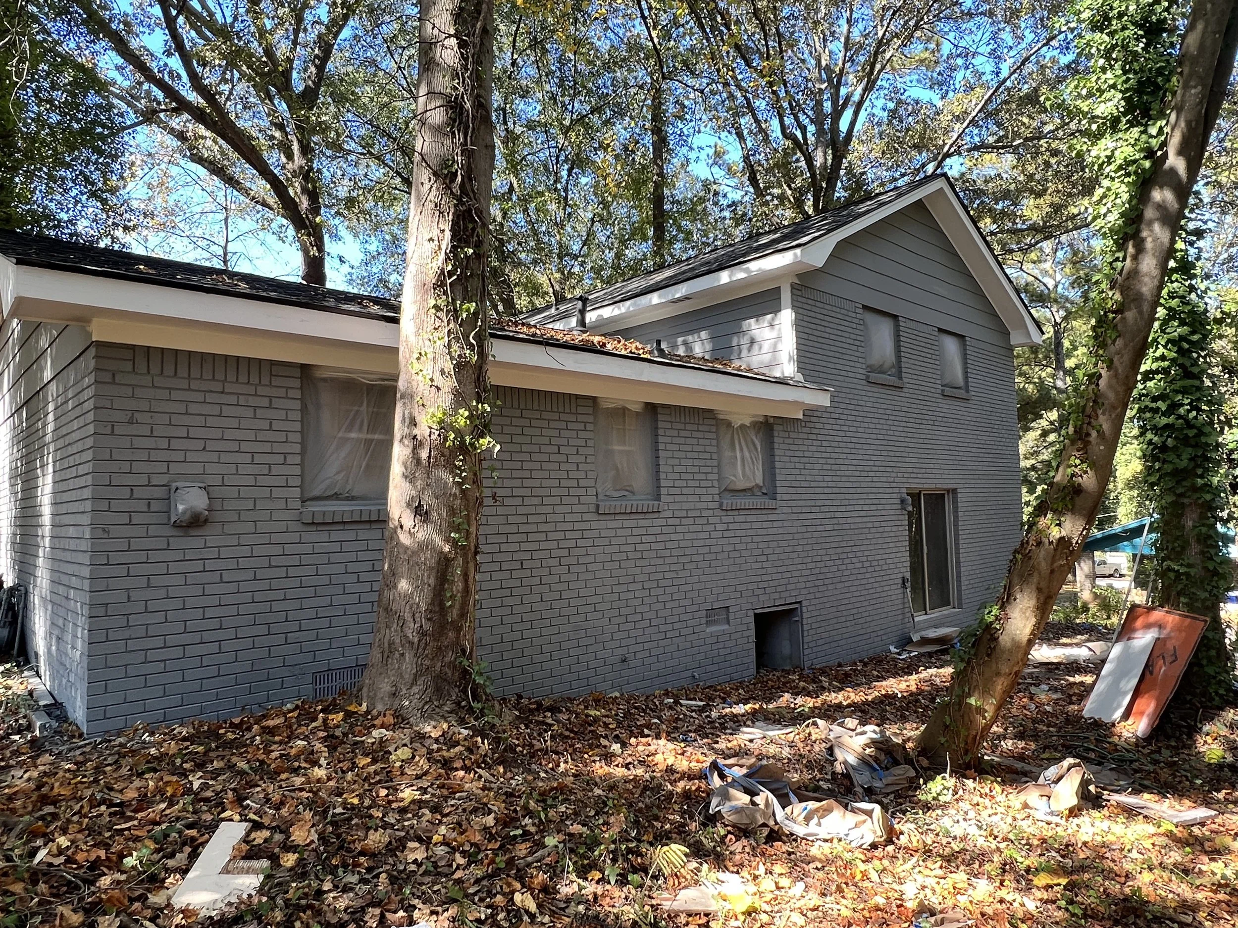 Side view of a two-story house under construction with gray brick exterior, surrounded by trees and fallen autumn leaves, with construction debris on the ground.