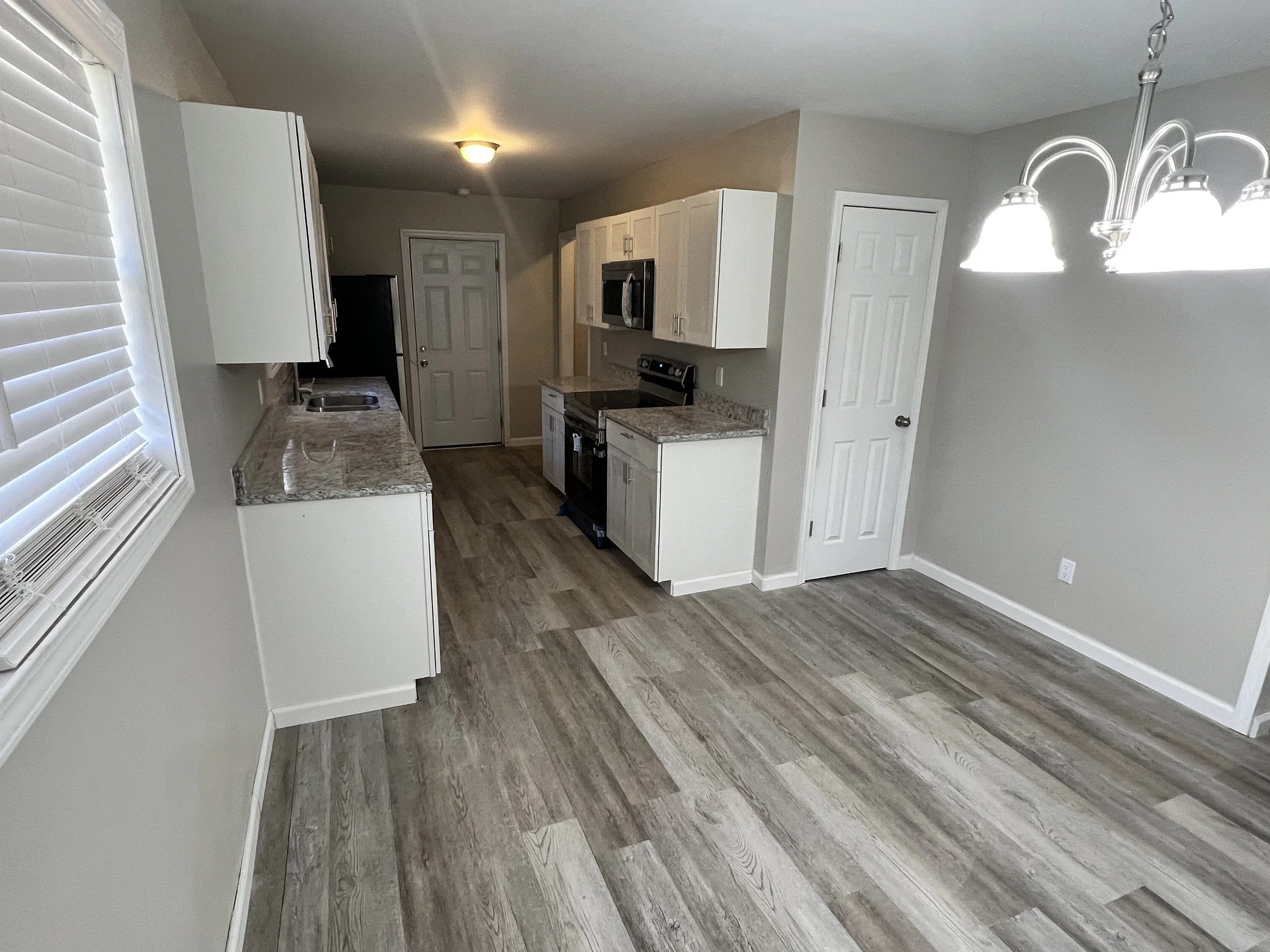 Empty kitchen with white cabinets, granite countertops, stainless steel appliances, a wooden floor, a window with blinds, and a chandelier lighting fixture.