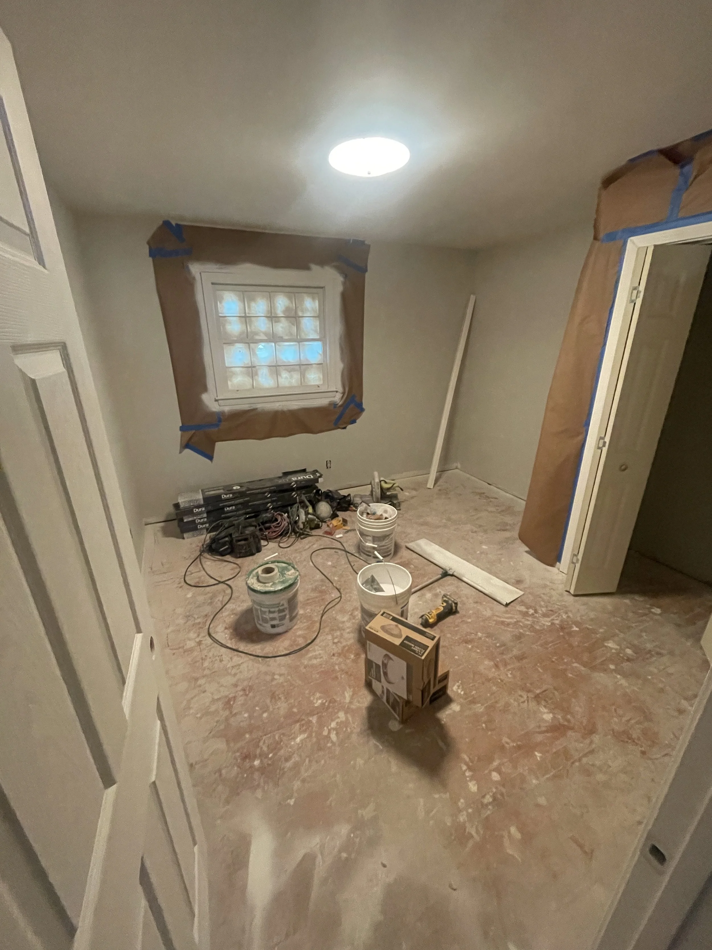A room under renovation with a window covered in brown paper and painter's tape, construction tools, paint buckets, and boxes scattered on the unfinished floor.