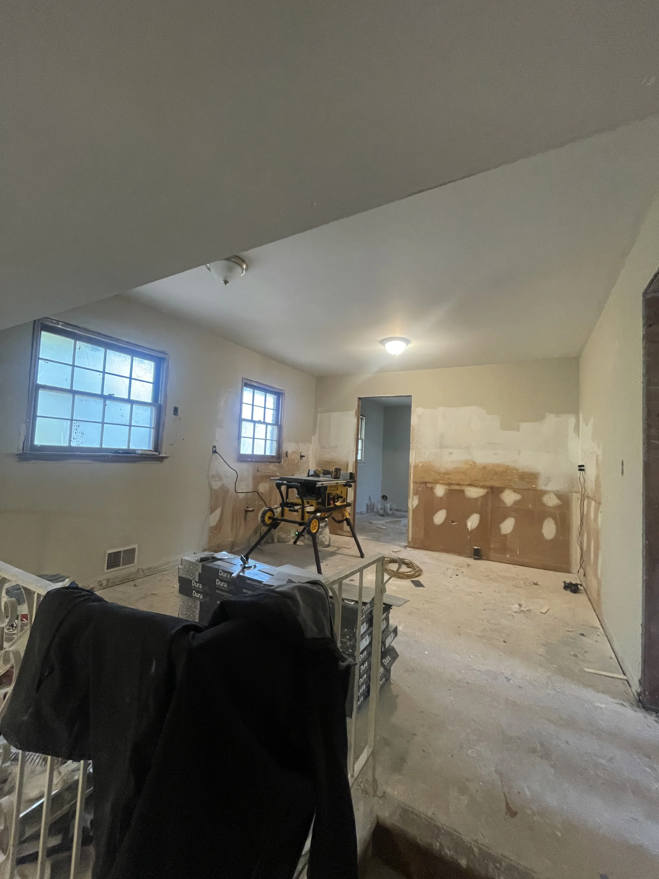 Room under construction with partially finished walls, two windows, construction tools and materials, and a doorway leading to another room.