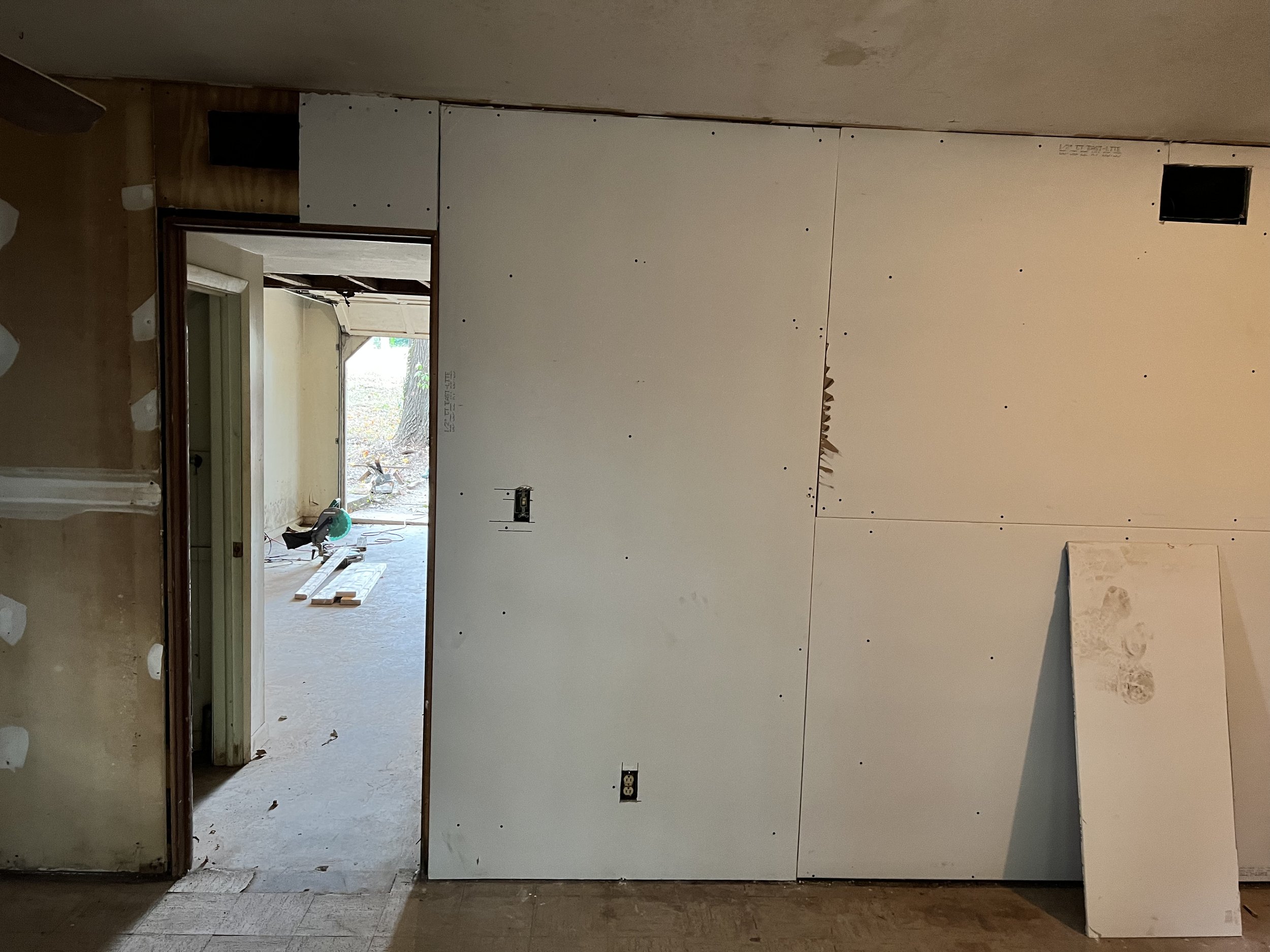 Interior view of a room under construction with drywall panels installed and electrical outlets visible, partially open doorway leading to an outdoor area with wooden planks and tools.